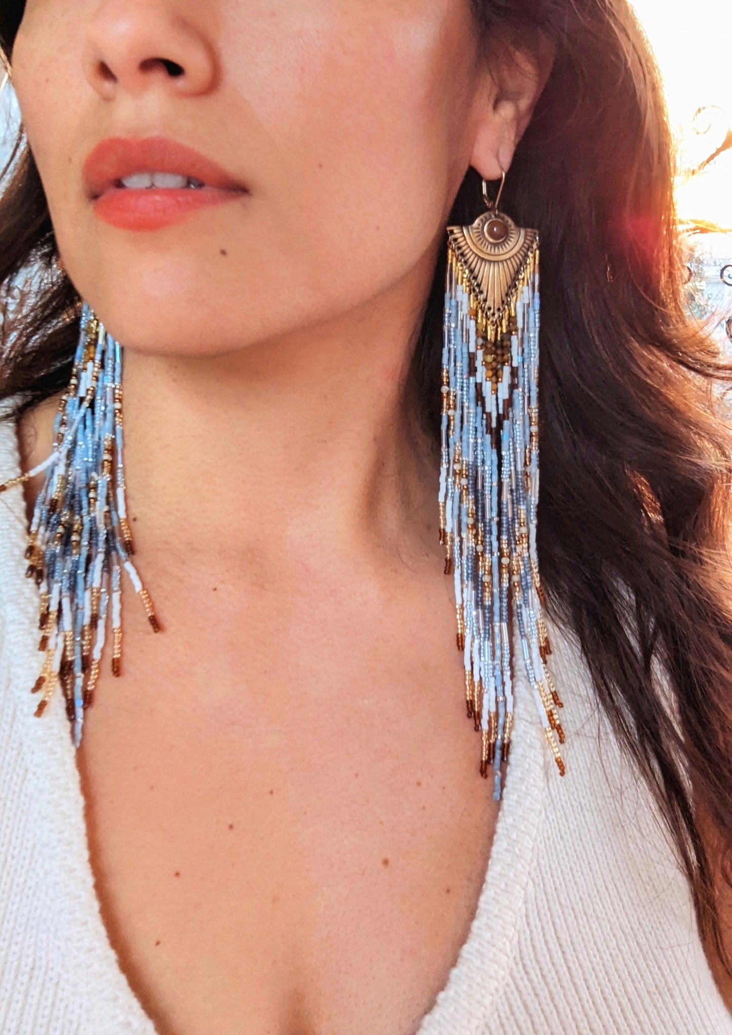 Woman wearing long dangling seed bead earrings with white, blue, and gold beads, featuring an ornate gold metal top. She has long dark hair.