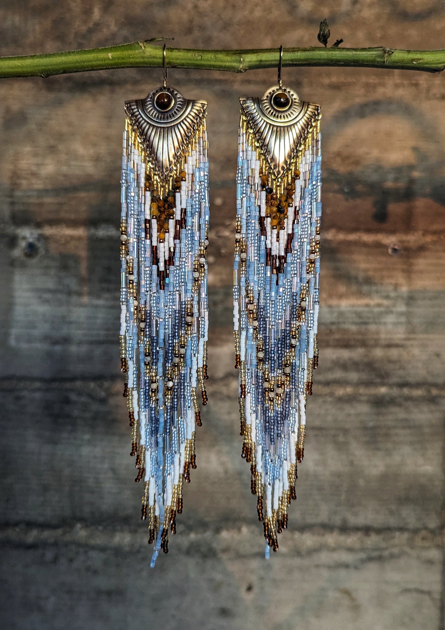 Long seed bead earrings with white, blue, and gold chevron patterns. Ornate gold metal tops. Hanging on a green bamboo stick.