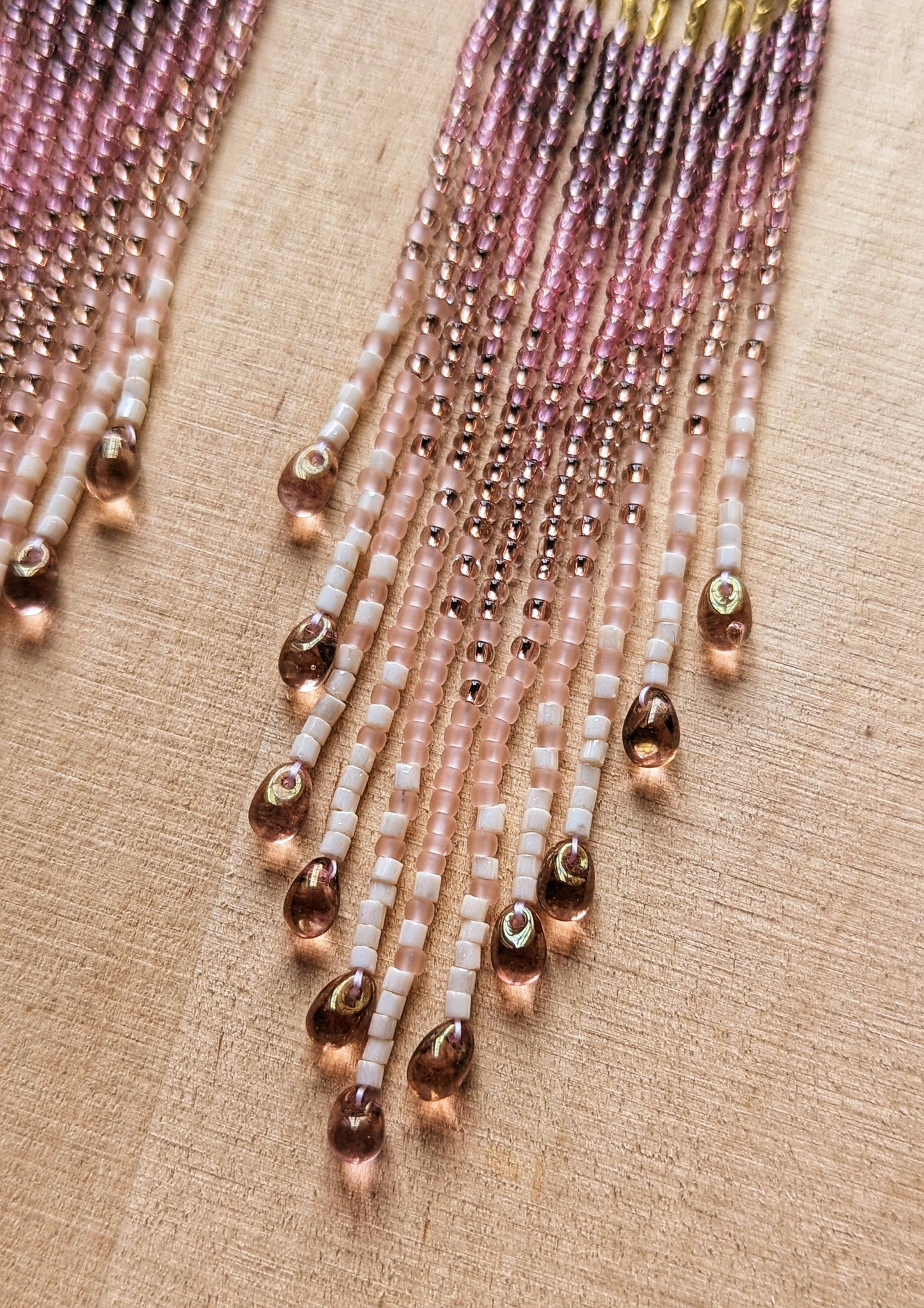 Handcrafted beaded fringe earrings featuring gold accents and soft pink hues, elegantly displayed on a wooden surface with a vibrant pink rose in the background.