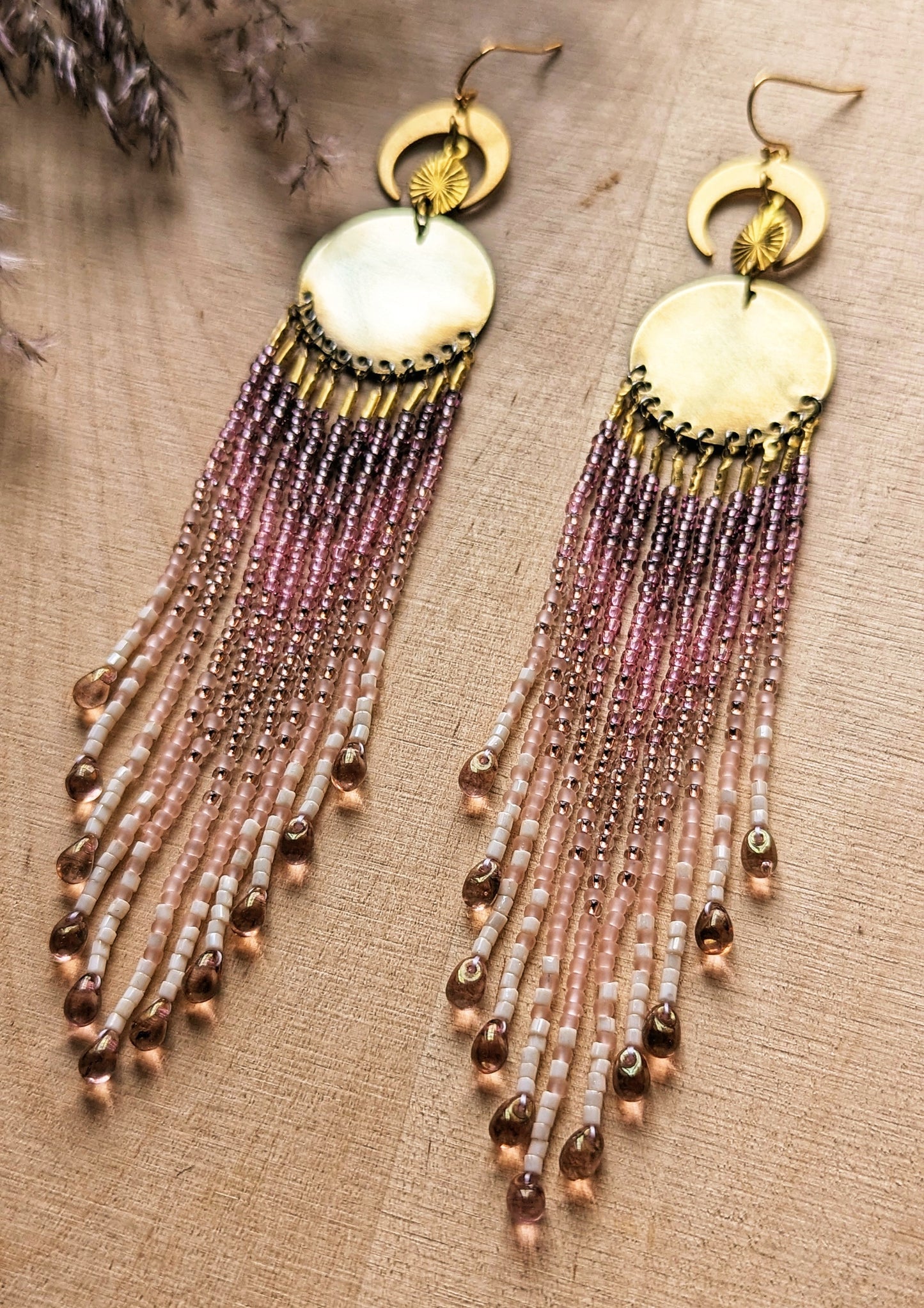 Handcrafted beaded fringe earrings featuring gold accents and soft pink hues, elegantly displayed on a wooden surface with a vibrant pink rose in the background.