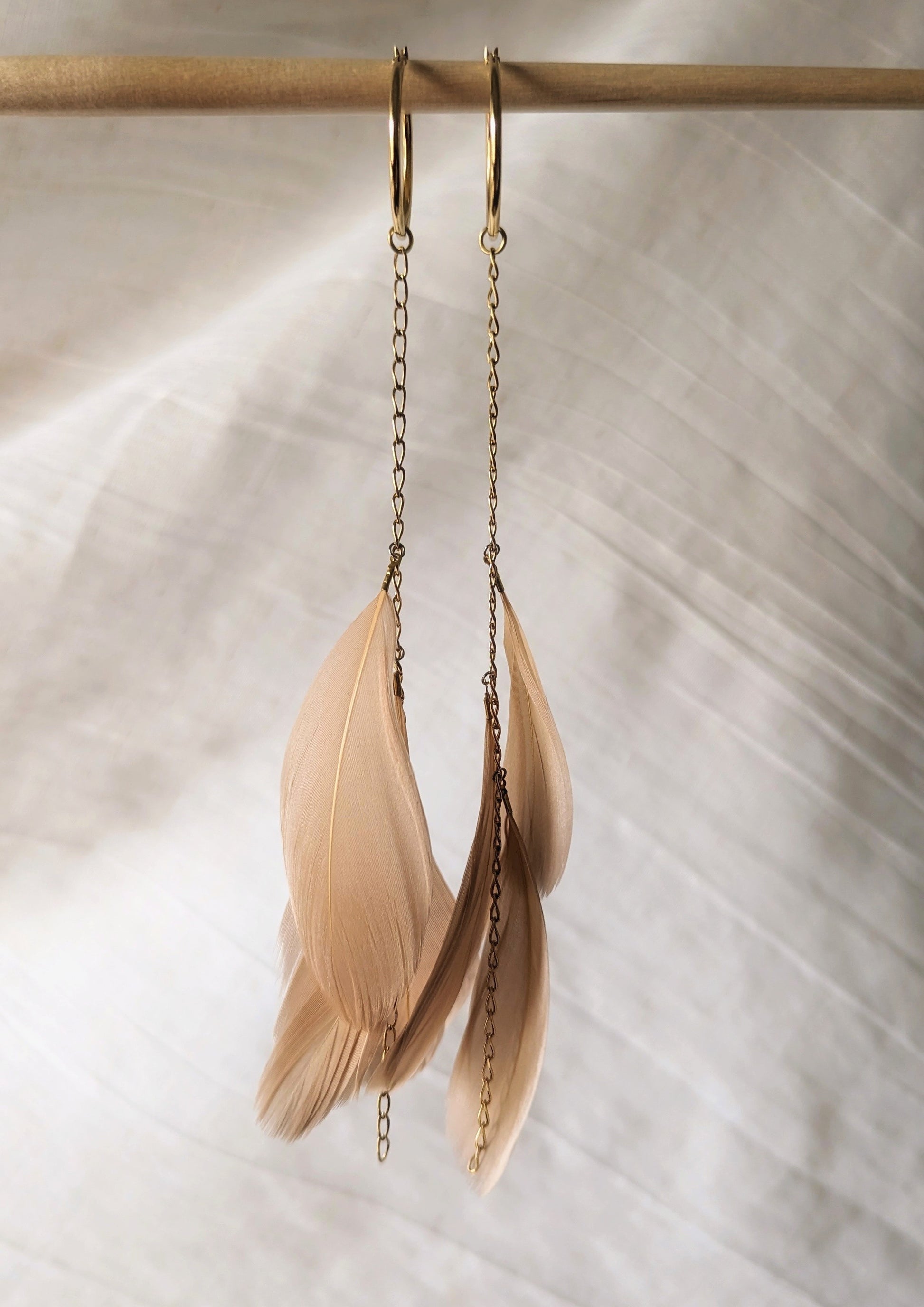 Blush feather earrings with gold hoop and chain detail from Moon & Milk, hanging against a soft white fabric backdrop.