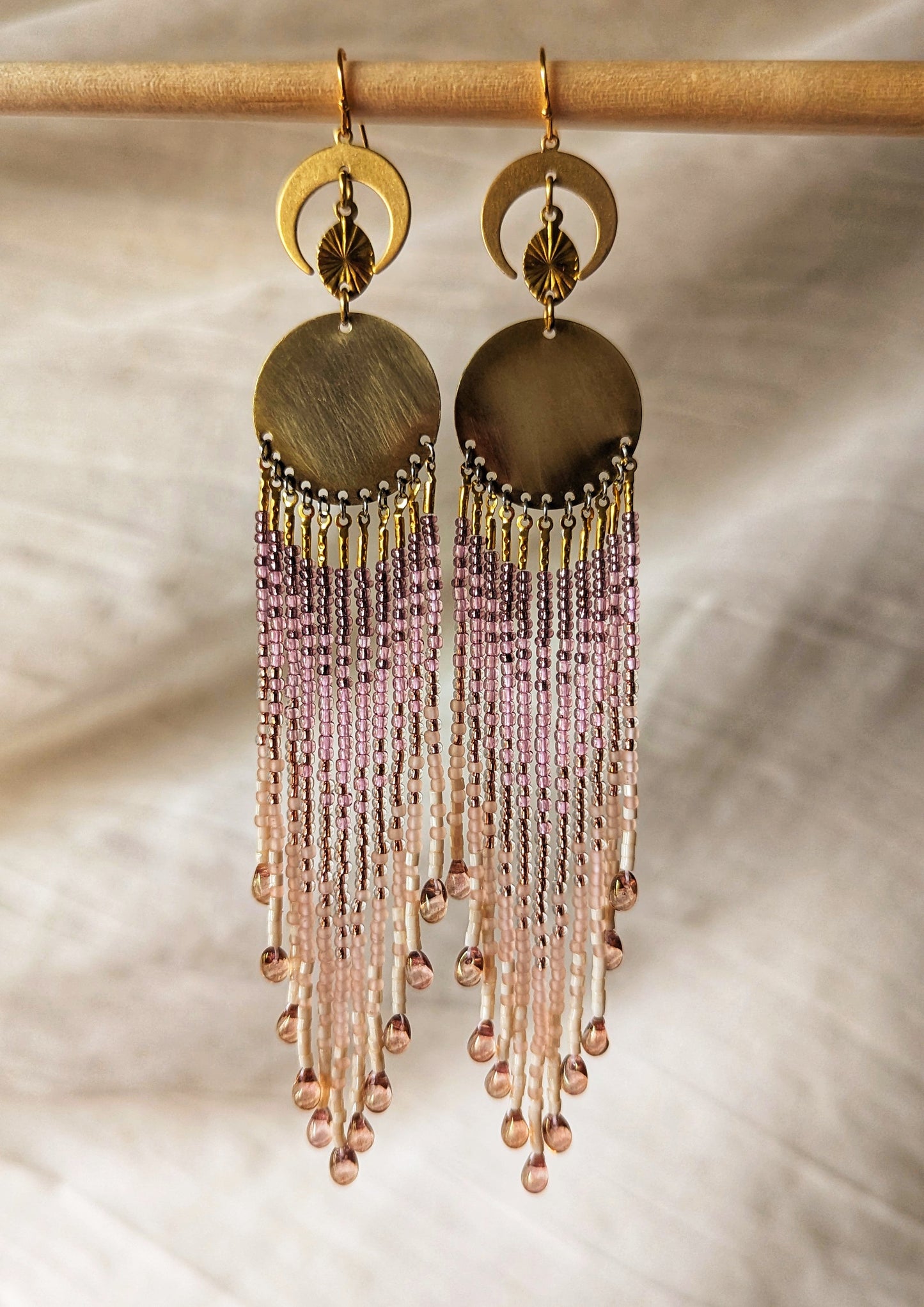 Intricate handcrafted earrings featuring cascading pink and lilac beads with crescent moon and circular brass details, elegantly hanging on a neutral backdrop.