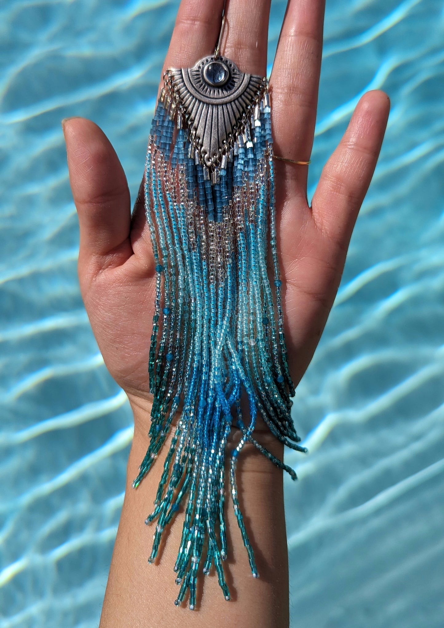 Moon & Milk - Stunning handmade beaded earrings with cascading blue and turquoise beads, featuring a silver-tone pendant with a central blue gemstone, elegantly displayed against a background of clear blue water.