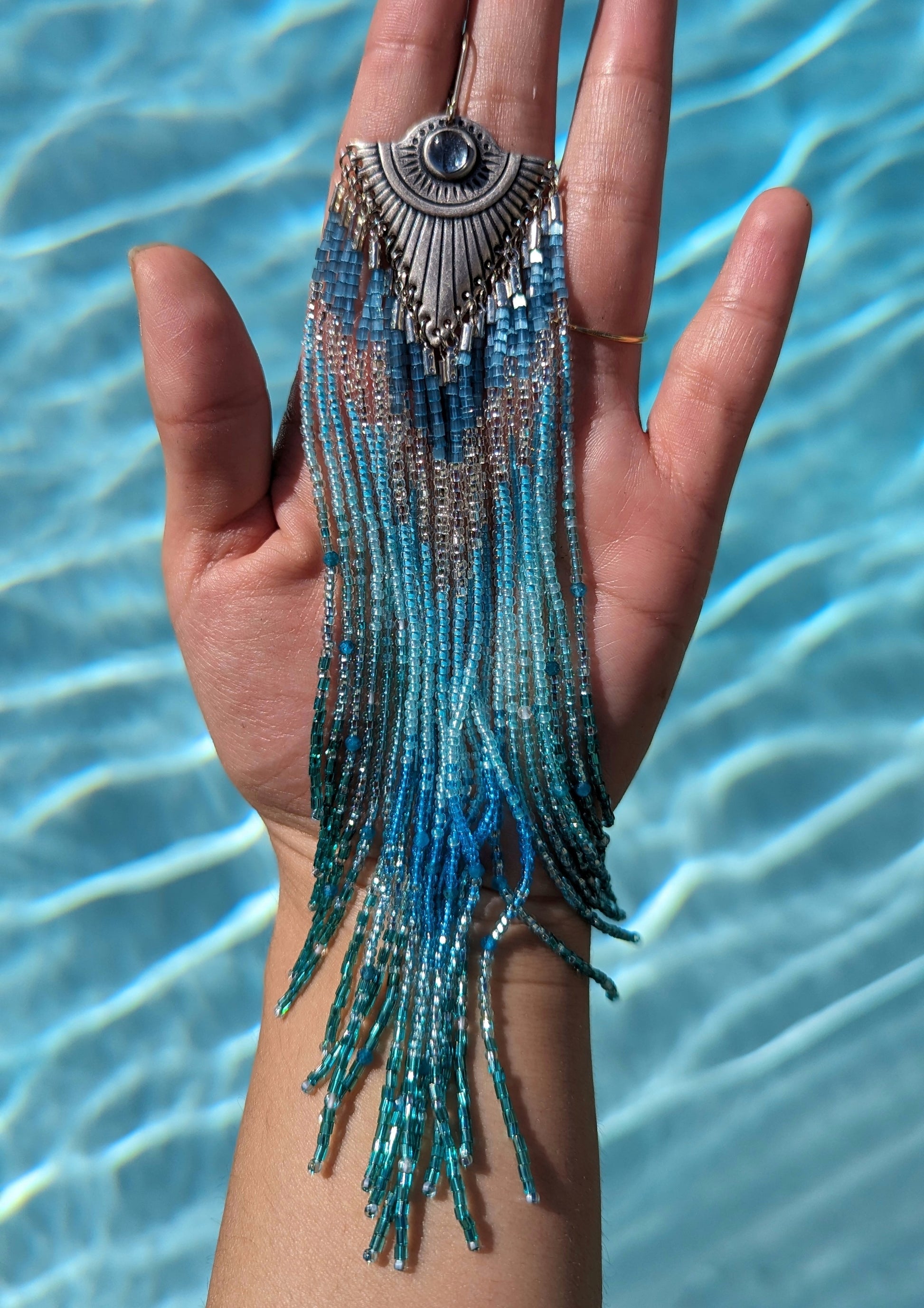 Moon & Milk - Stunning handmade beaded earrings with cascading blue and turquoise beads, featuring a silver-tone pendant with a central blue gemstone, elegantly displayed against a background of clear blue water.