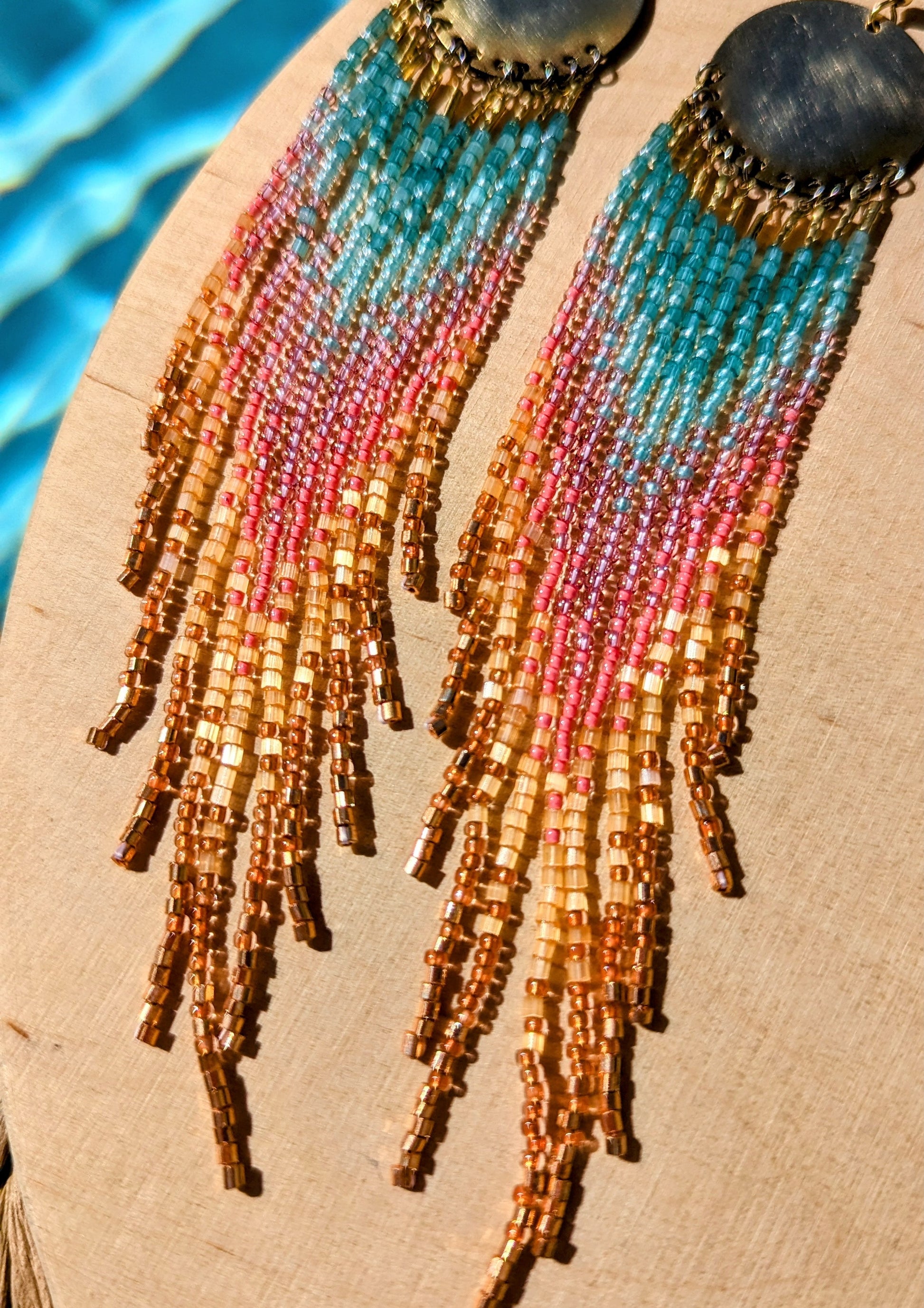 Handcrafted seed bead earrings with a vibrant ombre fringe design, transitioning from turquoise to pink to golden yellow, finished with copper-toned beads and a brass circular top.
