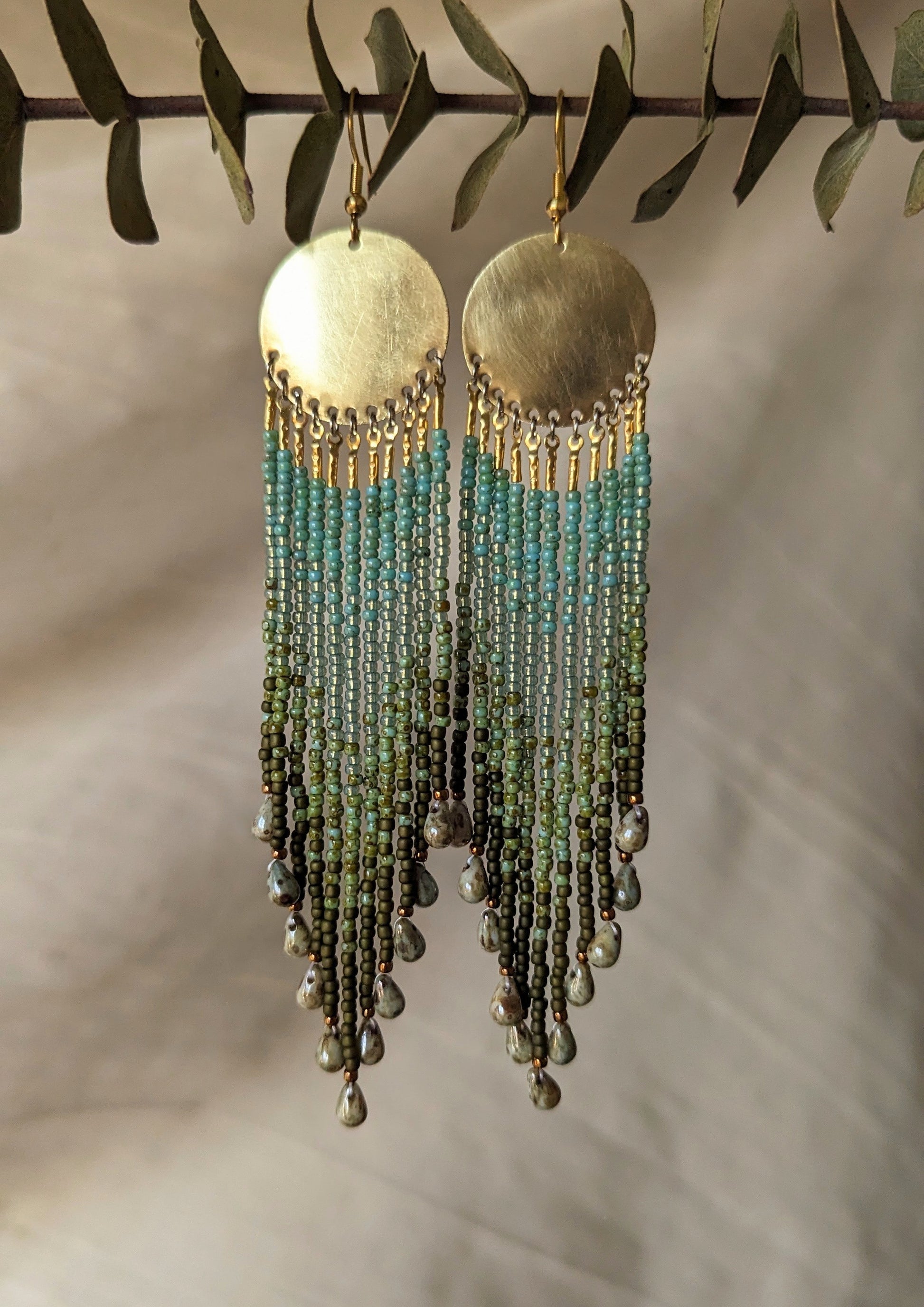 Moon & Milk long green ombré beaded earrings with brass circles and teardrop ends, handcrafted to echo forest textures and artisan beadwork style.