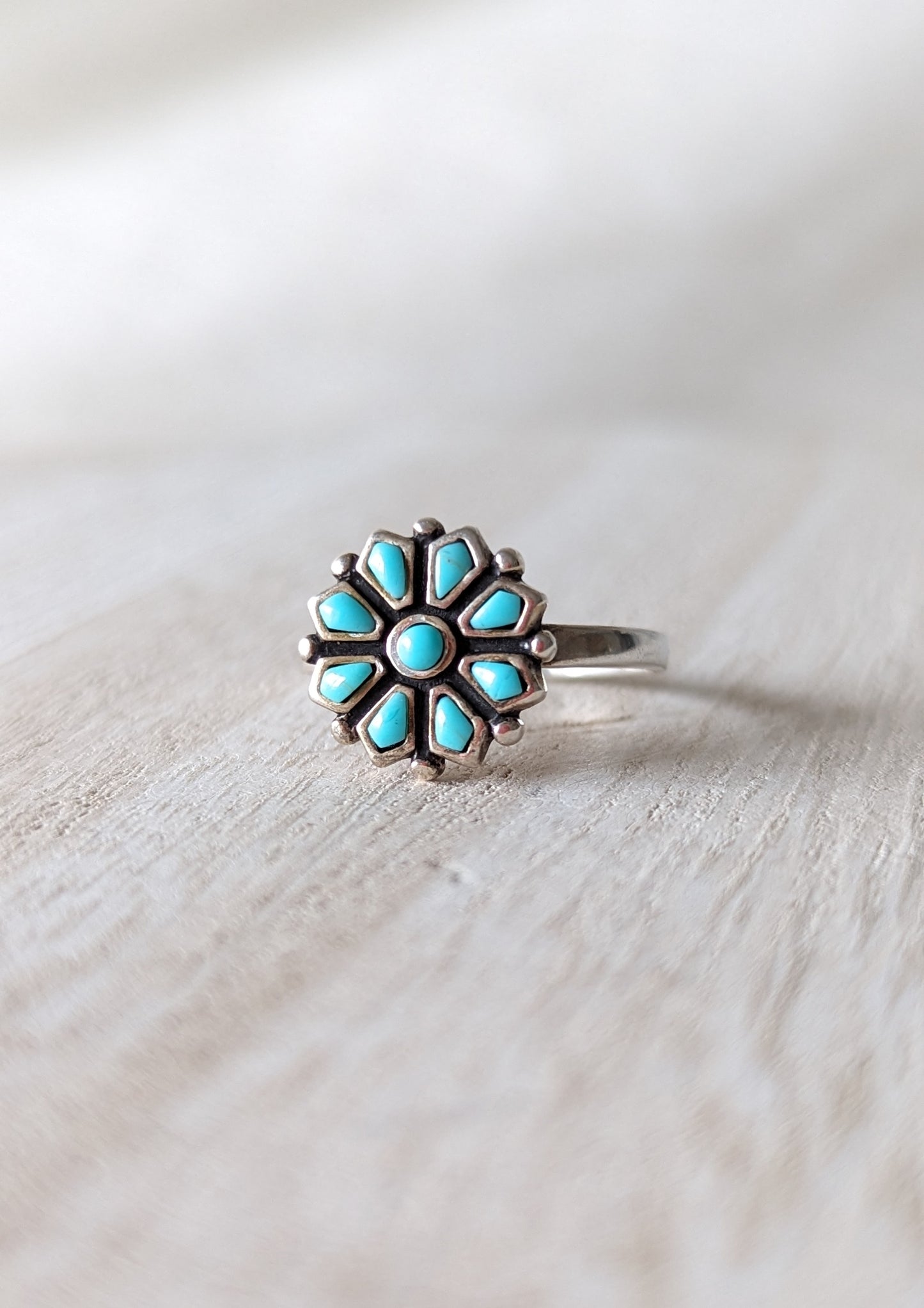 A sterling silver ring with a flower design, featuring turquoise gemstone petals. White rustic background.