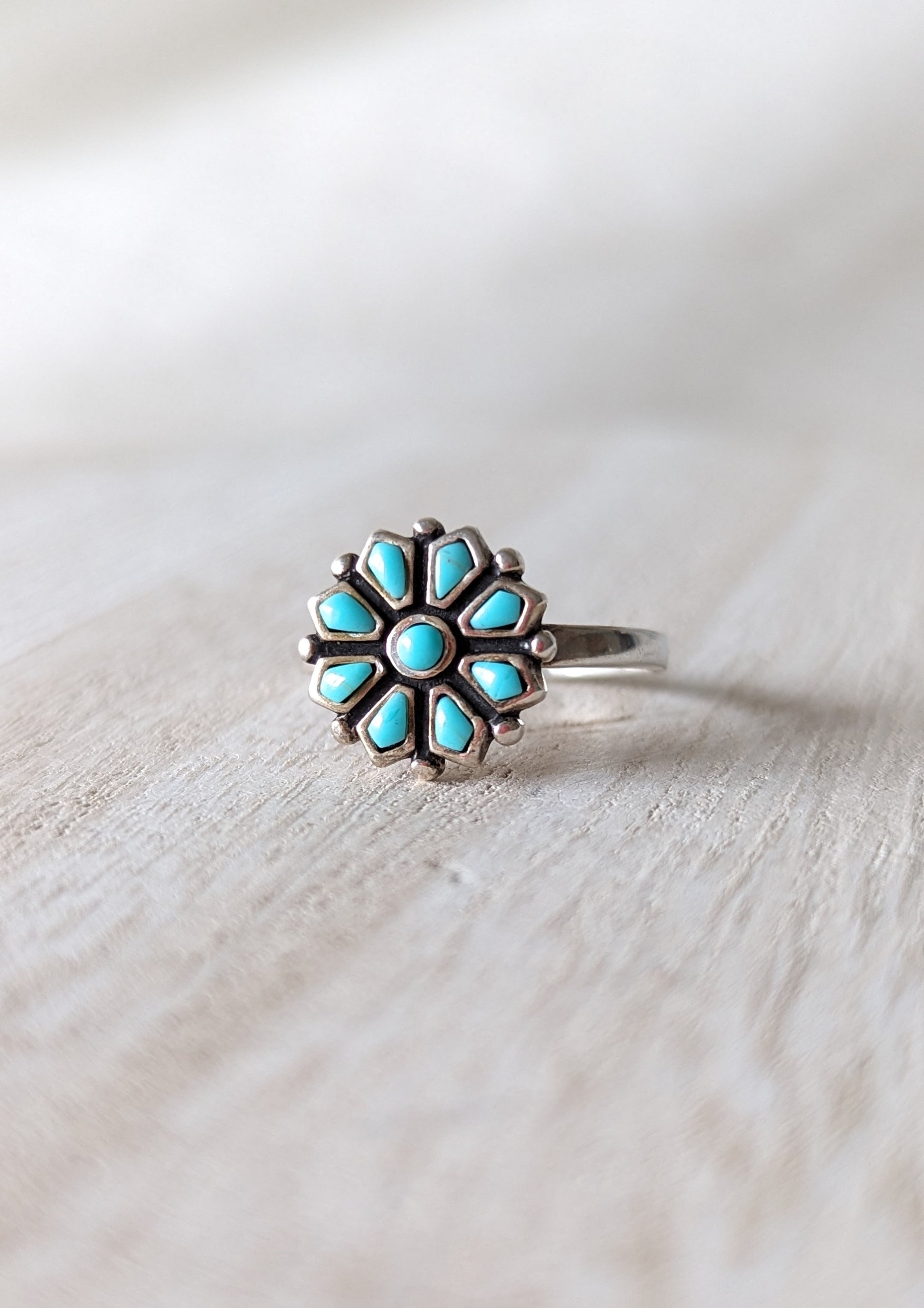A sterling silver ring with a flower design, featuring turquoise gemstone petals. White rustic background.