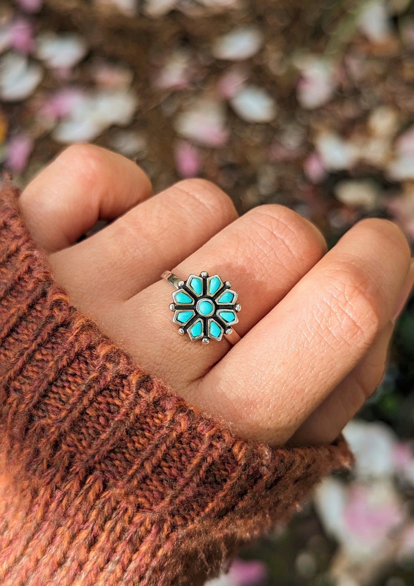 Hand wearing a sterling silver ring with a flower design, featuring turquoise gemstone petals. Background of blurred pink and white petals. Cozy brown sweater.