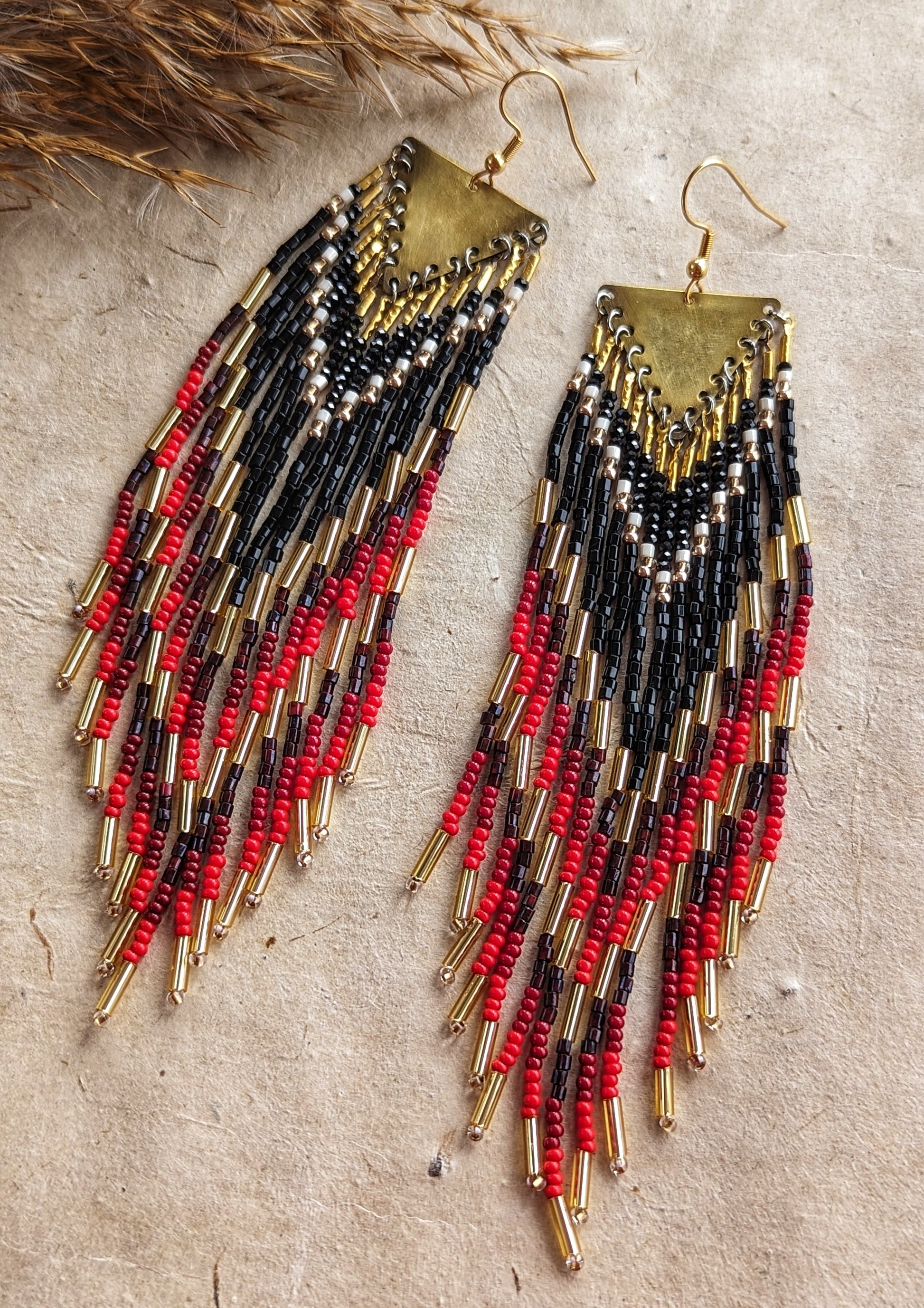 Moon & Milk earrings featuring black, red, and gold seed beads with brass triangle tops, laid on textured earth-toned fabric next to dried grasses.