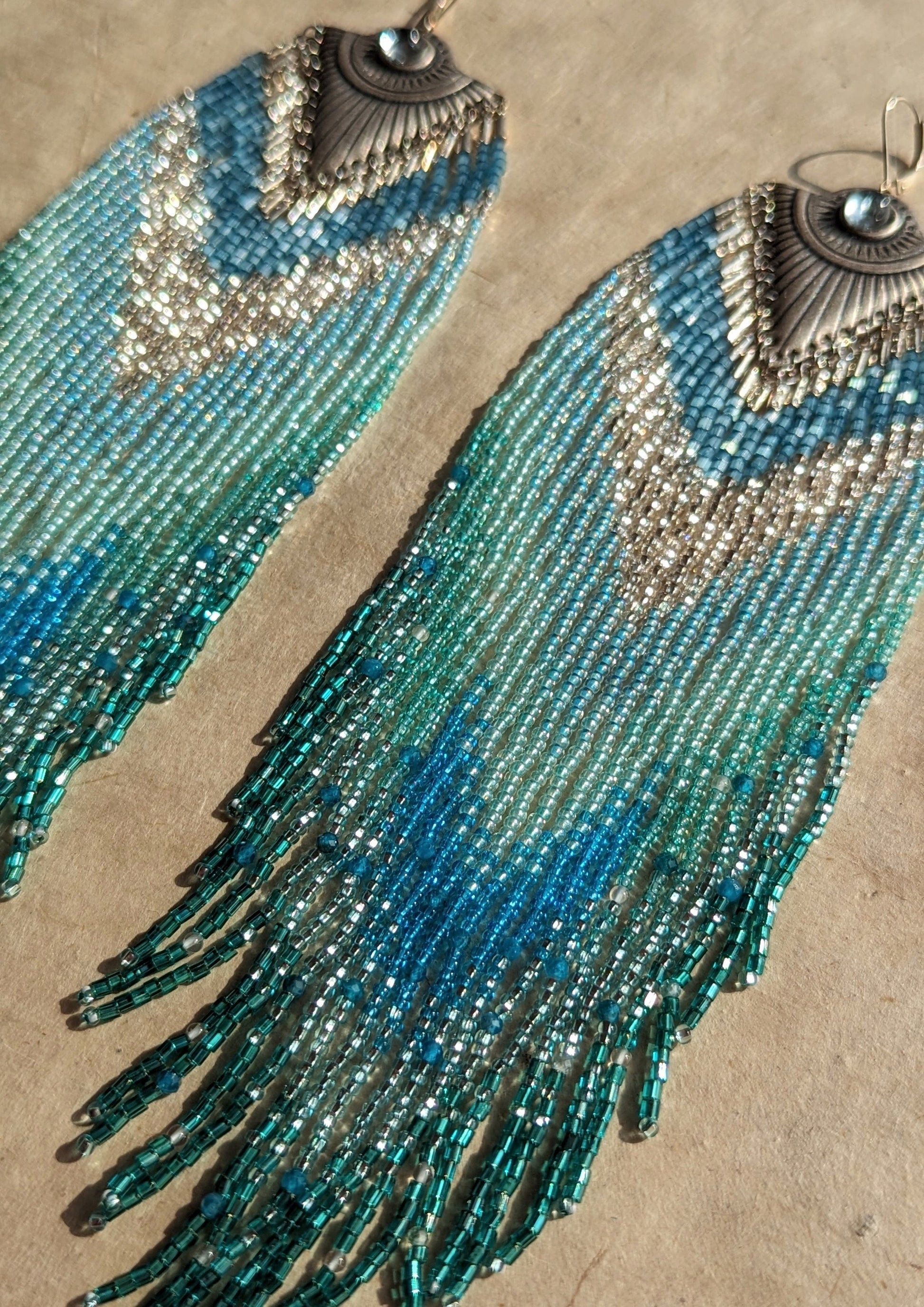 Moon & Milk - Handmade seed bead earrings featuring cascading blue and turquoise beads, accented with silver-tone pendants and central blue gemstones, elegantly displayed on a neutral background.