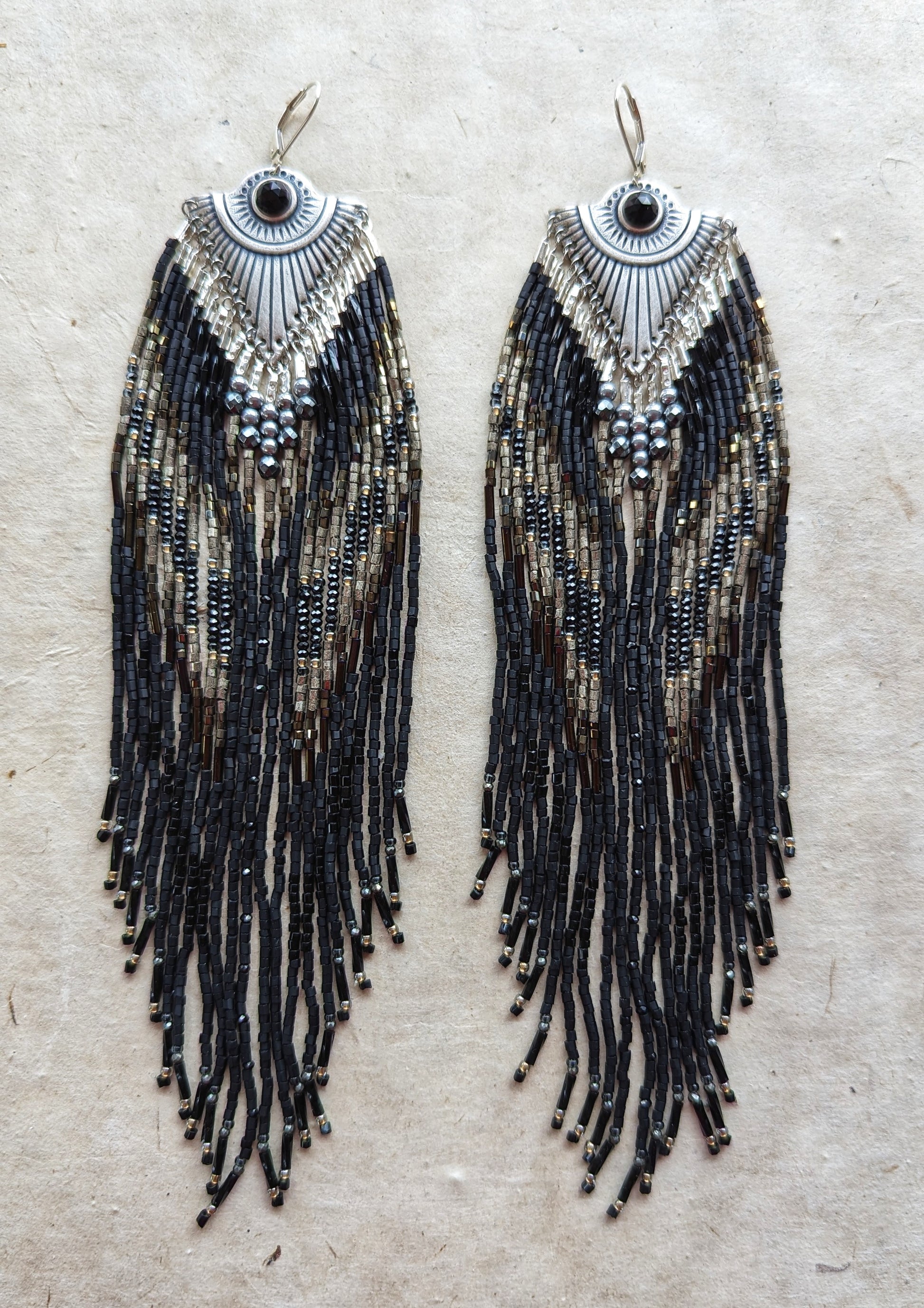 Handcrafted black beaded earrings with a fringe design. The beads feature a mix of black, silver, and gold tones, complemented by a silver fan-shaped top for an elegant, bold look