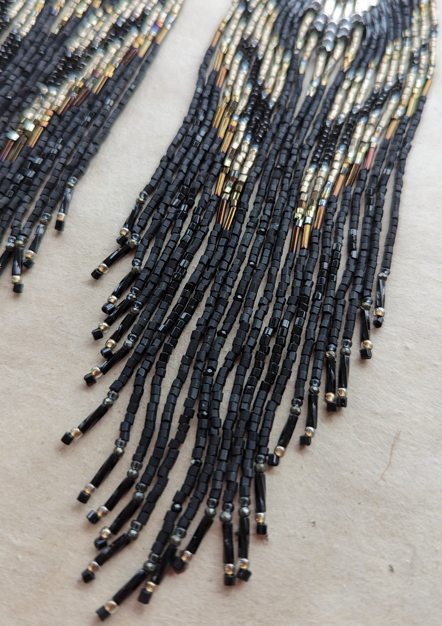 Handcrafted black beaded earrings with a fringe design. The beads feature a mix of black, silver, and gold tones, complemented by a silver fan-shaped top for an elegant, bold look