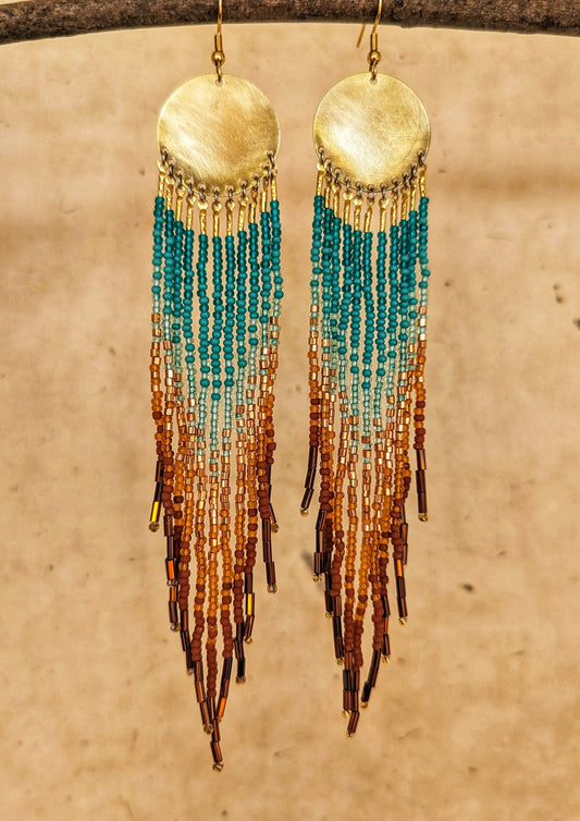 Handcrafted Moon & Milk earrings with circular brass tops and flowing strands of turquoise, amber, and brown seed beads against a warm beige background.