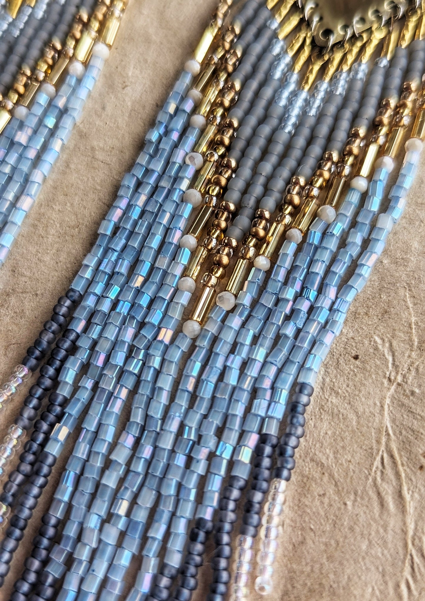 Moon & Milk beaded earrings inspired by Greek naiad Kyrene, featuring grey, pearl, and blue tones with long fringe on handcrafted brass triangles.