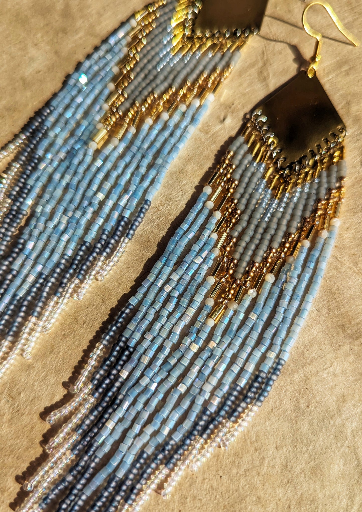 Moon & Milk beaded earrings inspired by Greek naiad Kyrene, featuring grey, pearl, and blue tones with long fringe on handcrafted brass triangles.