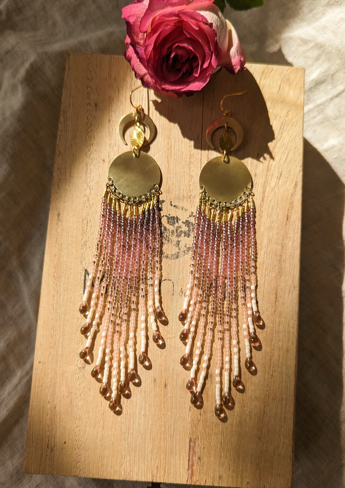 Handcrafted beaded fringe earrings featuring gold accents and soft pink hues, elegantly displayed on a wooden surface with a vibrant pink rose in the background.