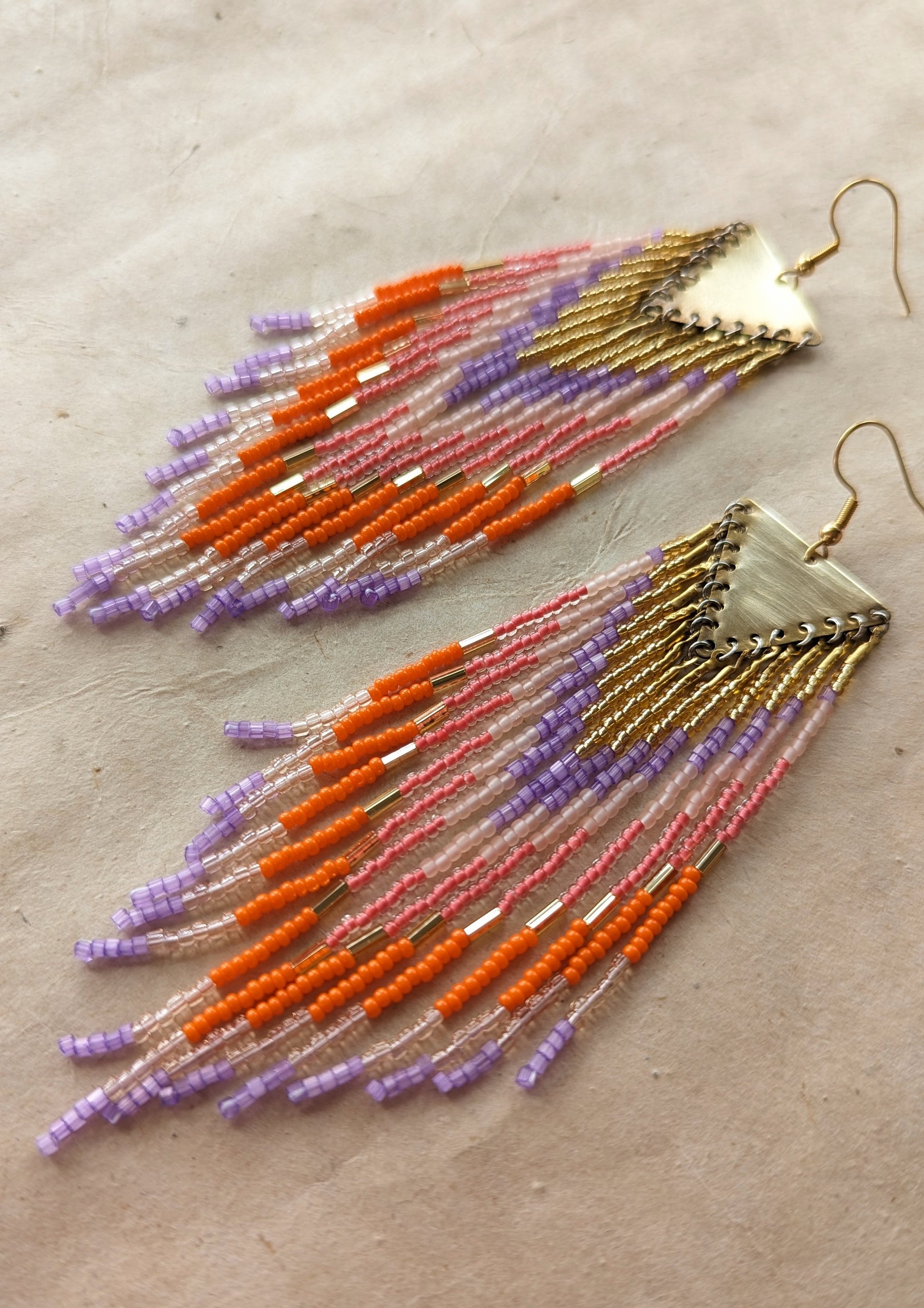Orange Popsicle earrings by Moon & Milk with orange, lavender, pink, and peach seed beads, handcrafted into long fringe hanging from brass triangle tops.
