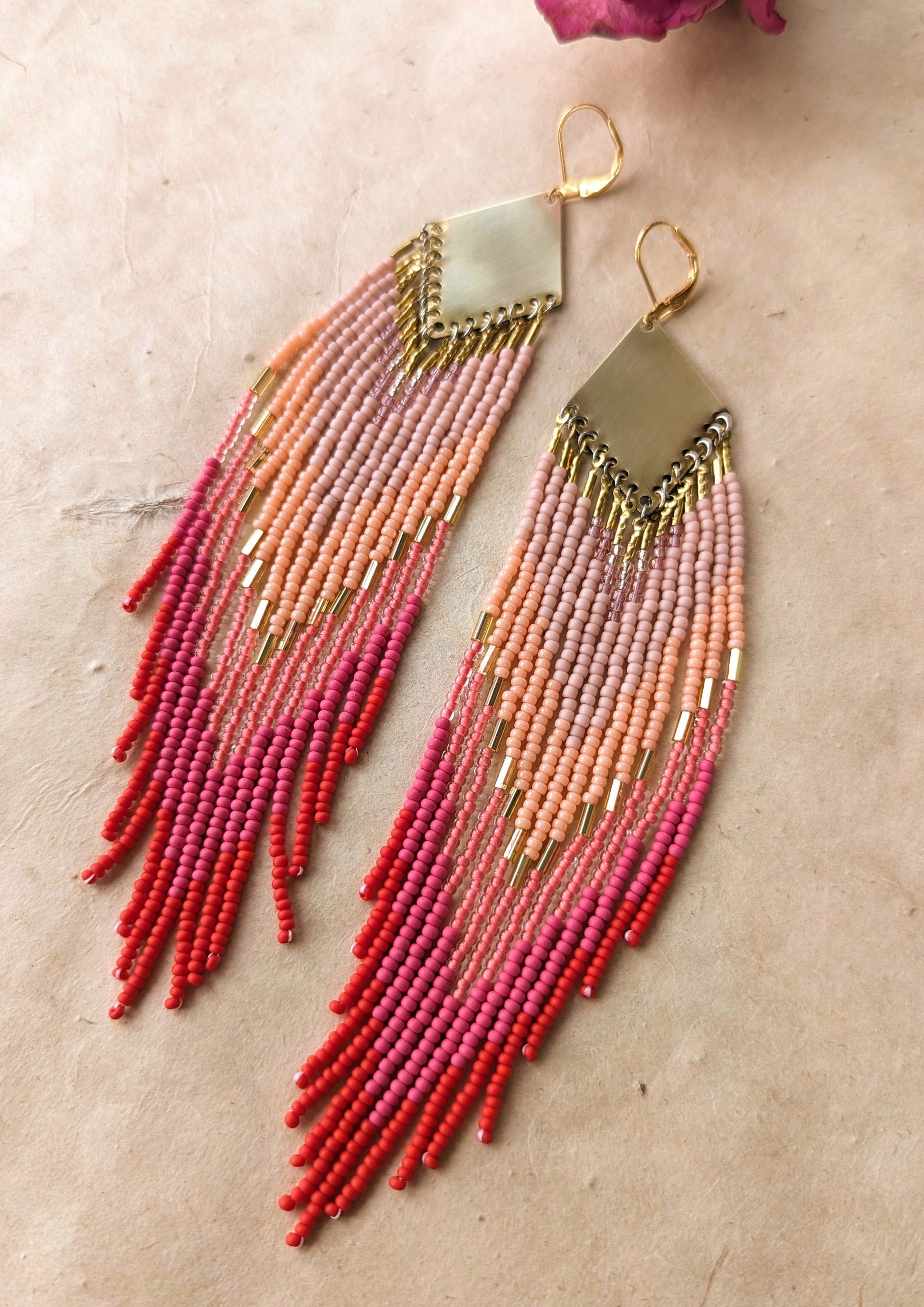 Moon & Milk handcrafted beaded earrings in ombré shades of rose, coral, and red with brass findings, displayed beside a dried pink rose on linen.