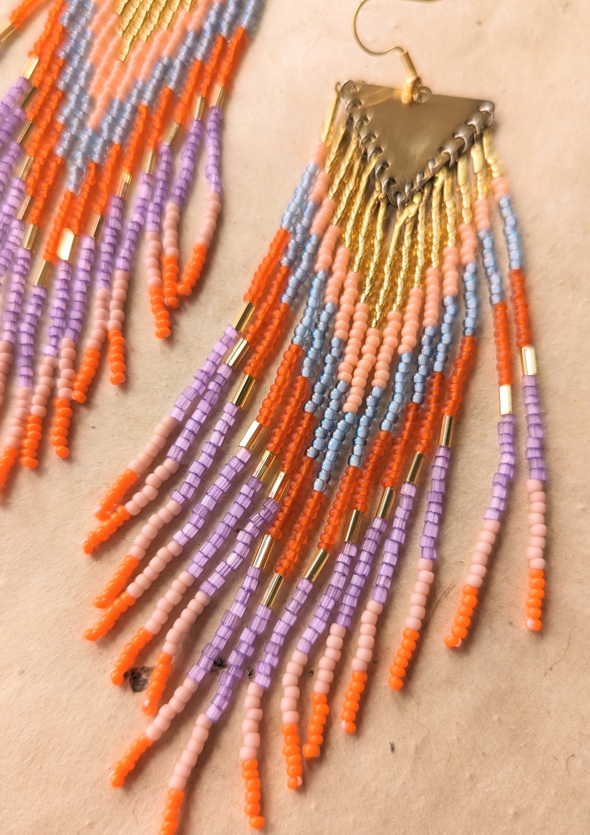 Colorful seed bead earrings with shades of orange, pink, purple,  gold, and light blue arranged in a triangular design, hanging from a brass finding.