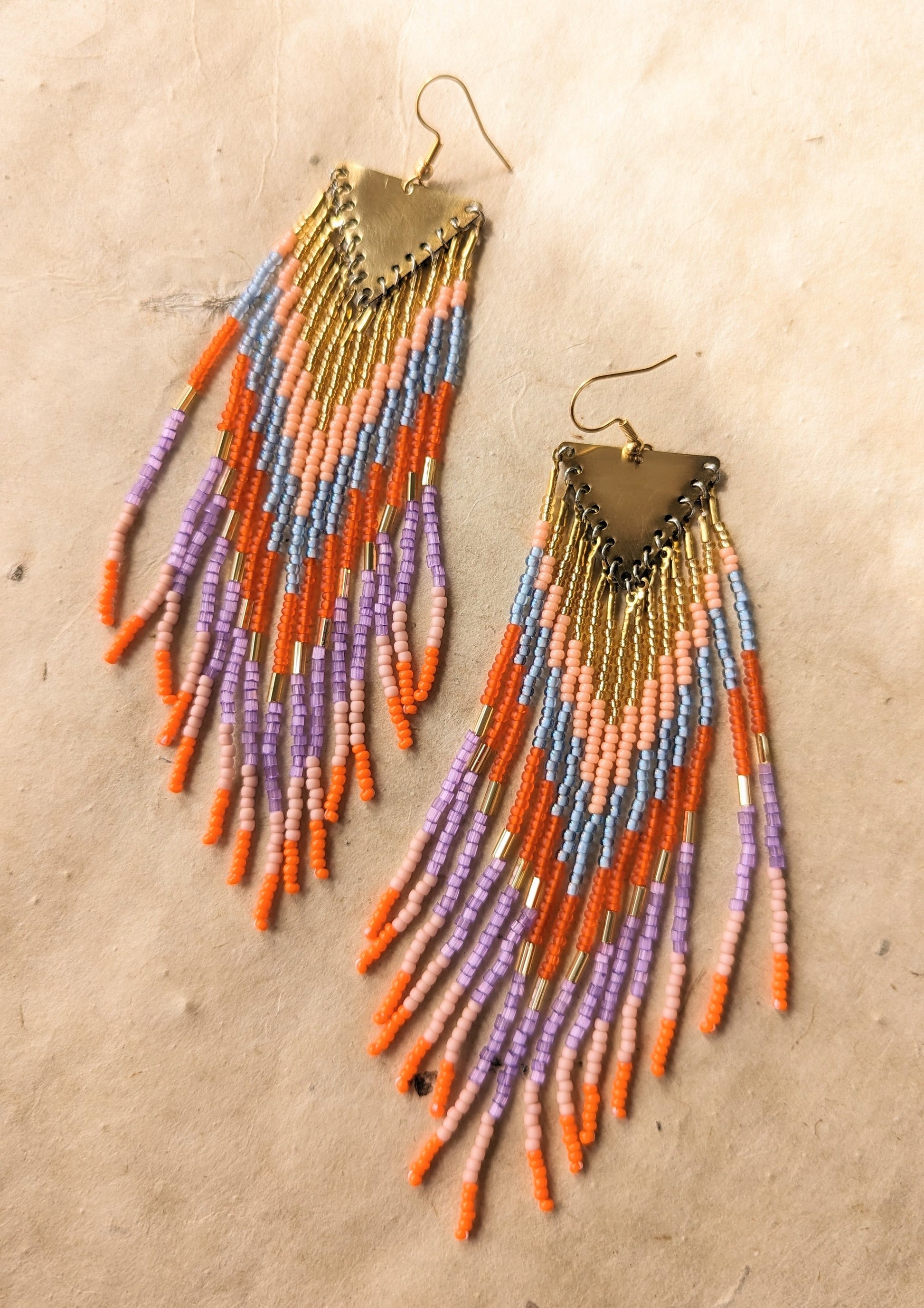 Colorful seed bead earrings with shades of orange, pink, purple,  gold, and light blue arranged in a triangular design, hanging from a brass finding.