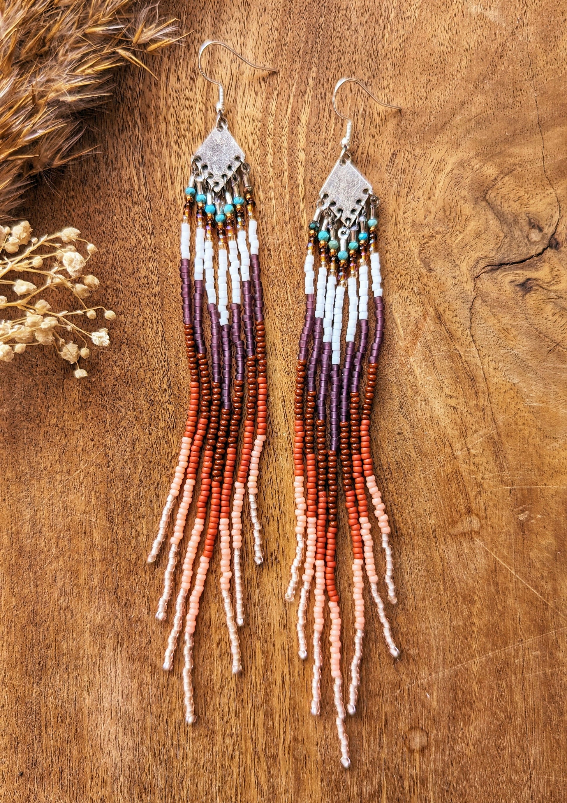 Long beaded fringe earrings with earthy tones of brown, terracotta, and white, displayed on a wooden background with dried floral accents.-Moon & Milk