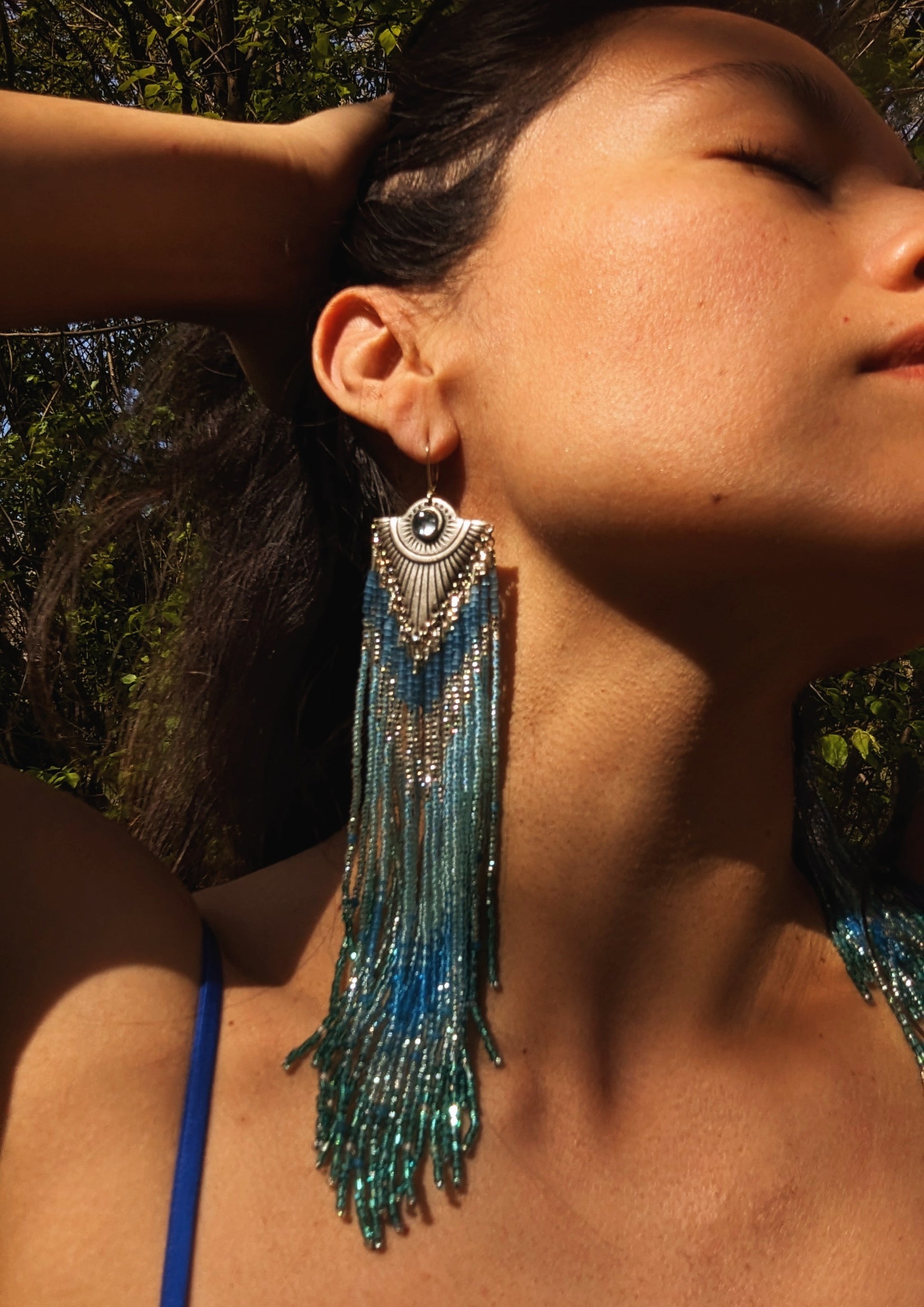 Moon & Milk - Woman wearing handmade beaded earrings with cascading blue and turquoise beads, featuring a silver-tone pendant with a central blue gemstone, beautifully highlighted by natural sunlight.