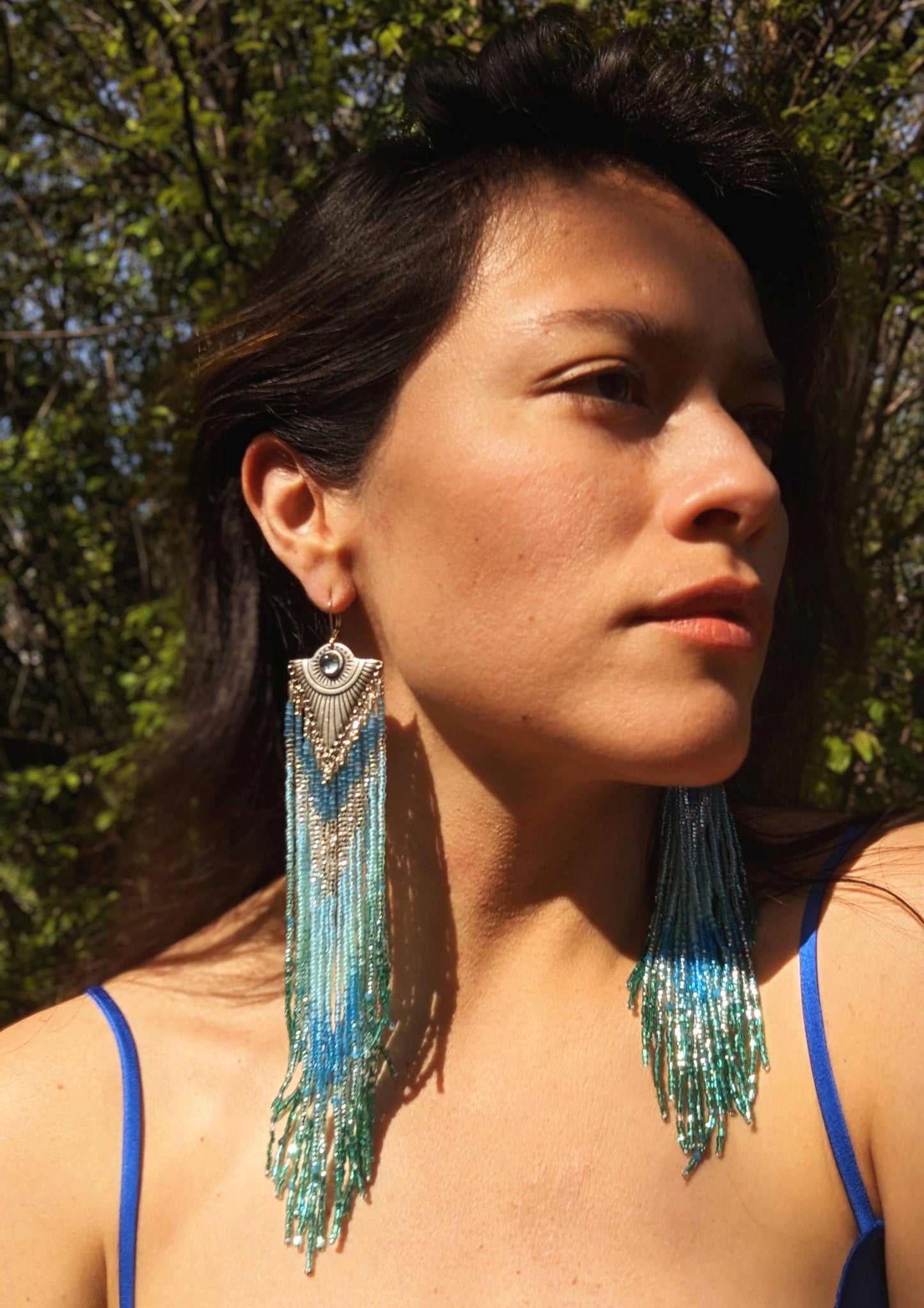 Moon & Milk - Woman wearing handmade beaded earrings with cascading blue and turquoise beads, featuring a silver-tone pendant with a central blue gemstone, beautifully highlighted by natural sunlight.