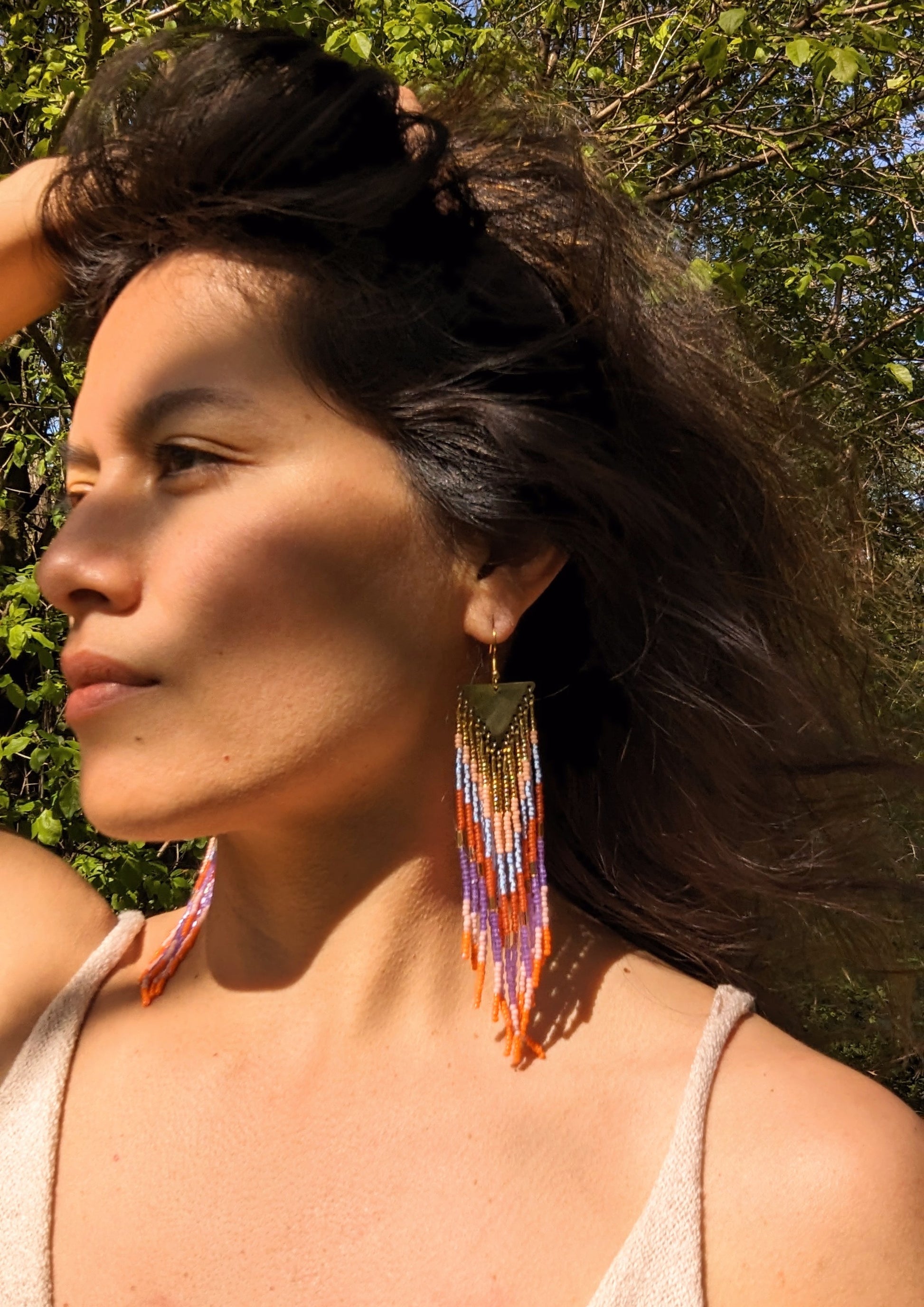 Woman wearing long, colorful beaded earrings in orange, pink, purple, and blue hues, accented by brass, with wind blowing through her hair in a natural outdoor setting.
