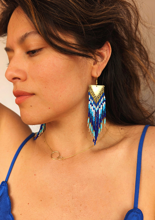 Woman wearing handcrafted  native earrings with cobalt, turquoise, and white beadwork, brass findings, and fringe design. Inspired by Utah landscapes.