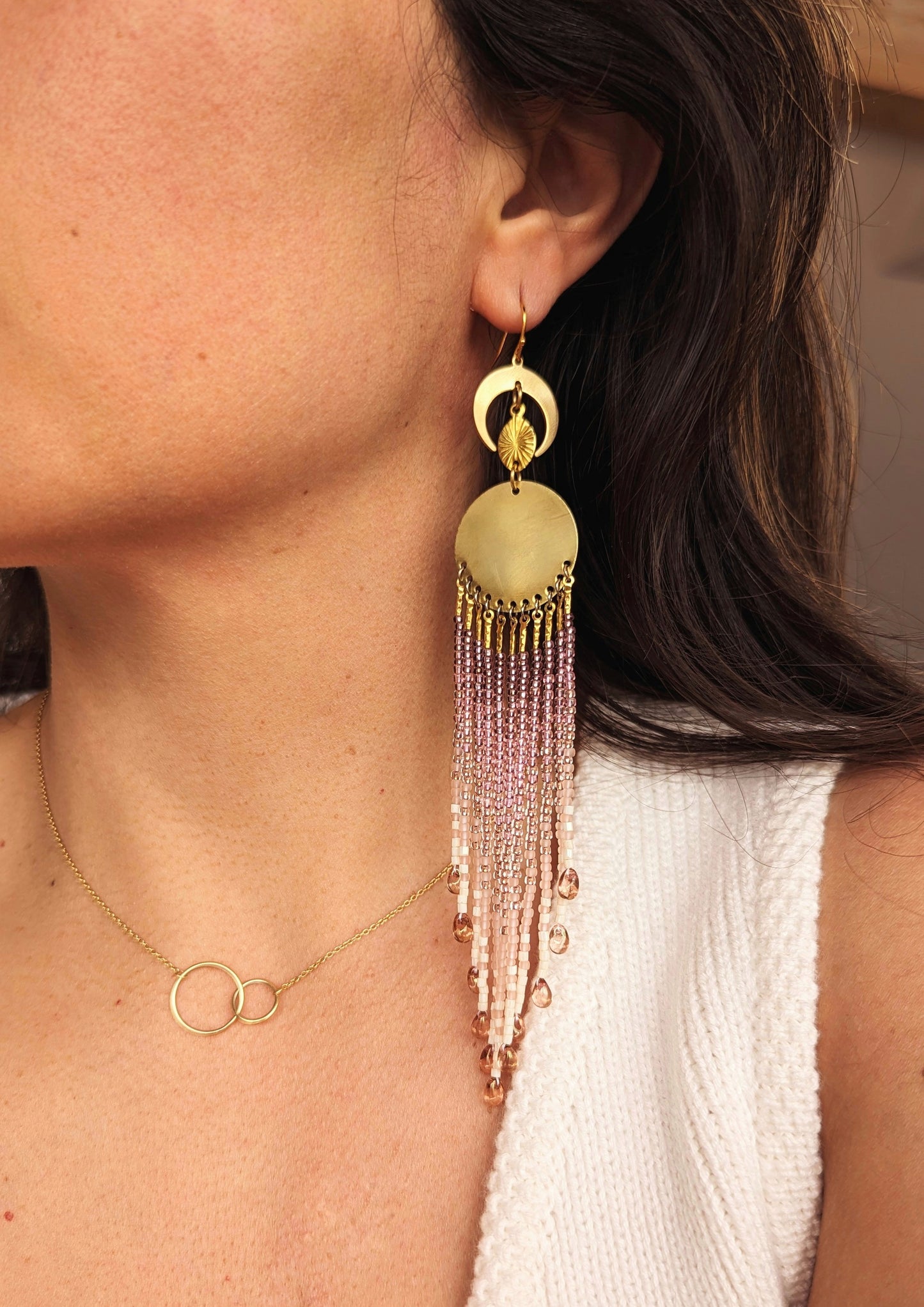 Close-up of a woman wearing elegant handcrafted beaded fringe earrings with cascading pink and gold beads, crescent moon, and brass details, paired with a minimalist gold necklace.