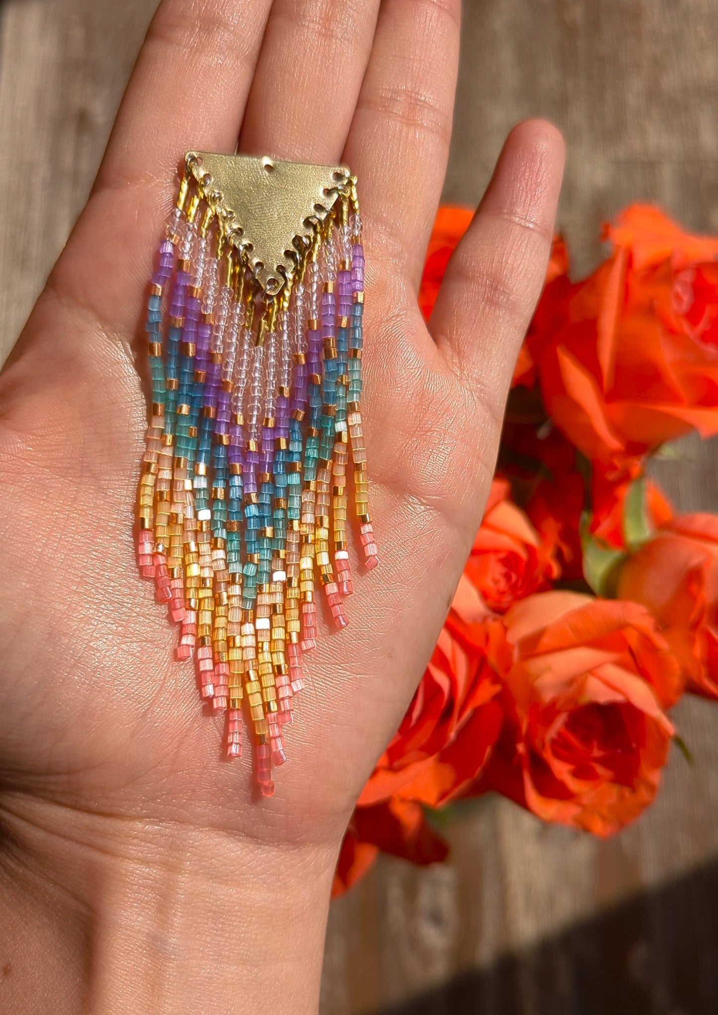 Moon & Milk rainbow beaded earring with brass triangle top, displayed on a hand with vibrant orange roses in the background.