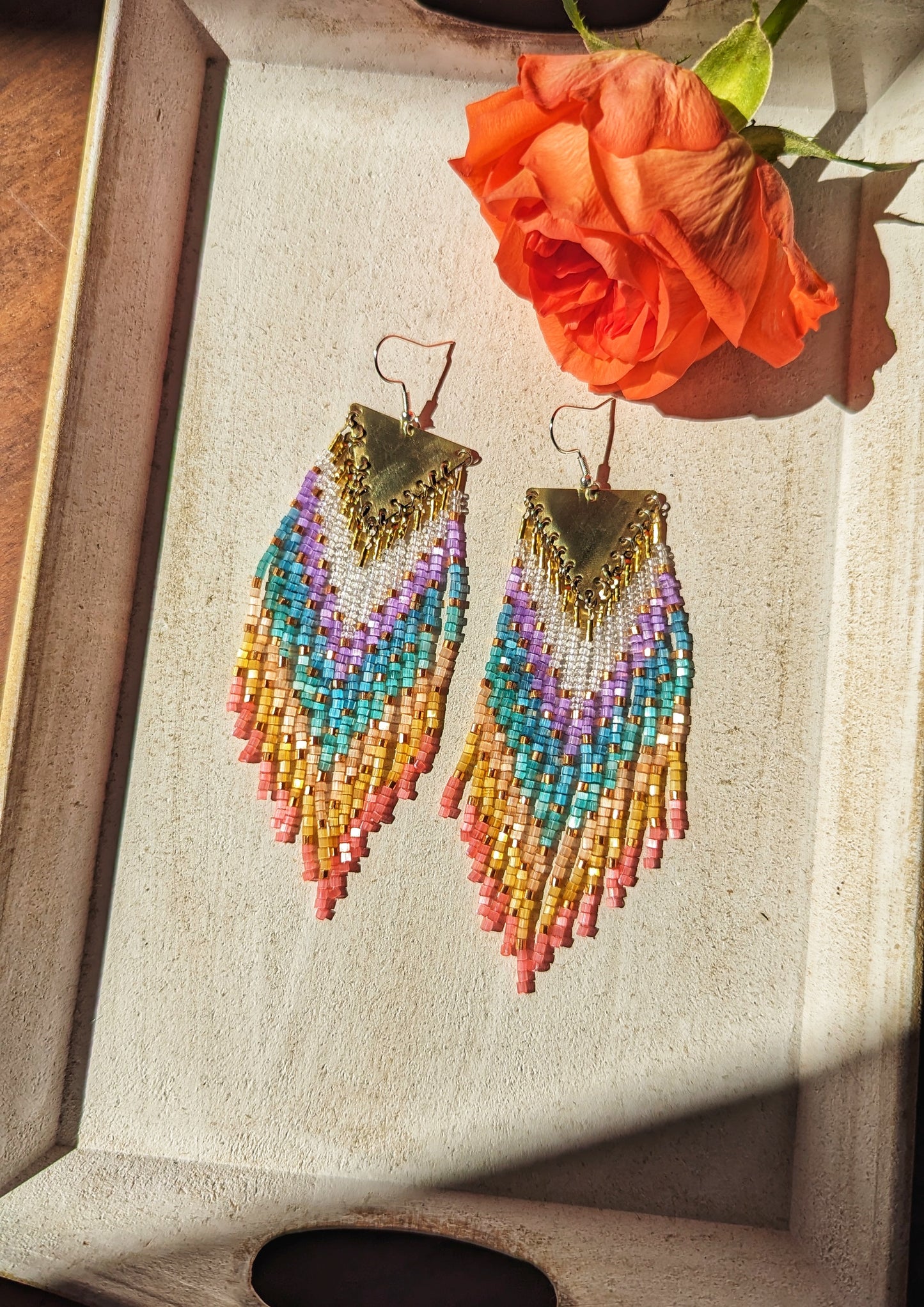 Moon & Milk rainbow beaded earring with brass triangle top, displayed on a hand with vibrant orange roses in the background.