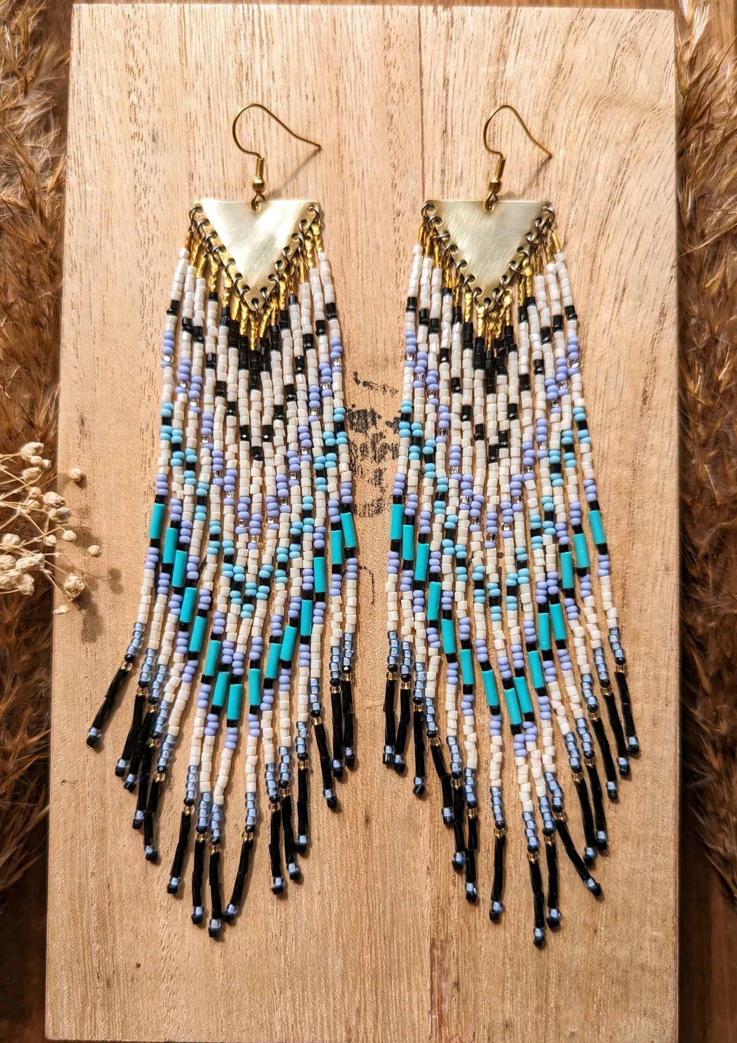 Moon & Milk long native beaded earrings with white, black, icy blue, and turquoise beadwork on brass triangles, displayed on wood.