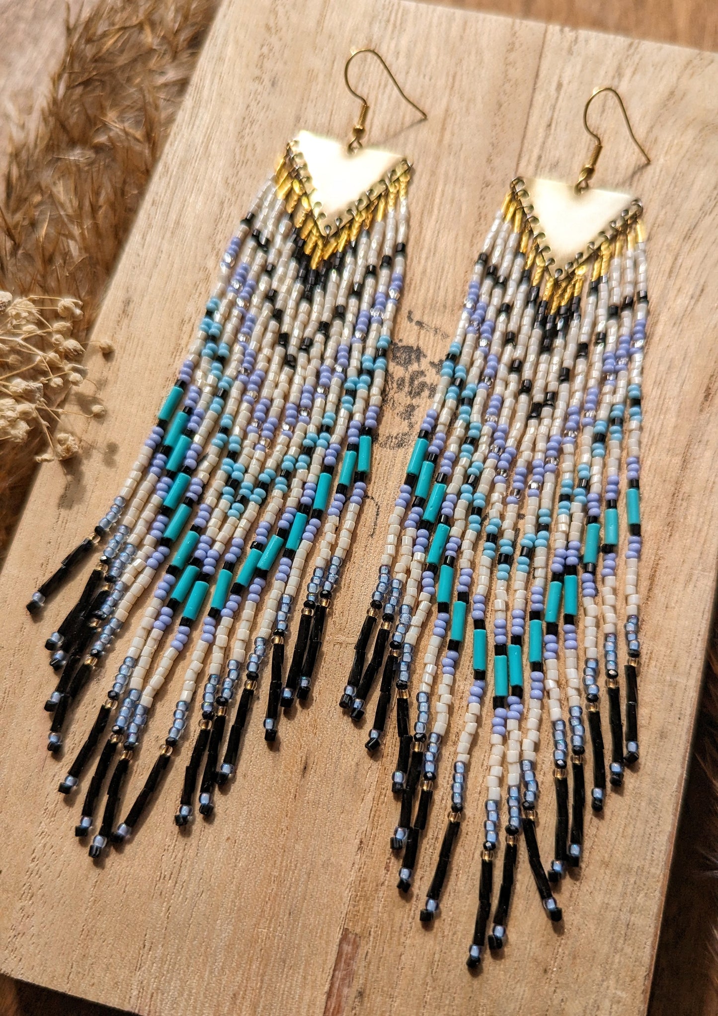 Moon & Milk long native beaded earrings with white, black, icy blue, and turquoise beadwork on brass triangles, displayed on wood.