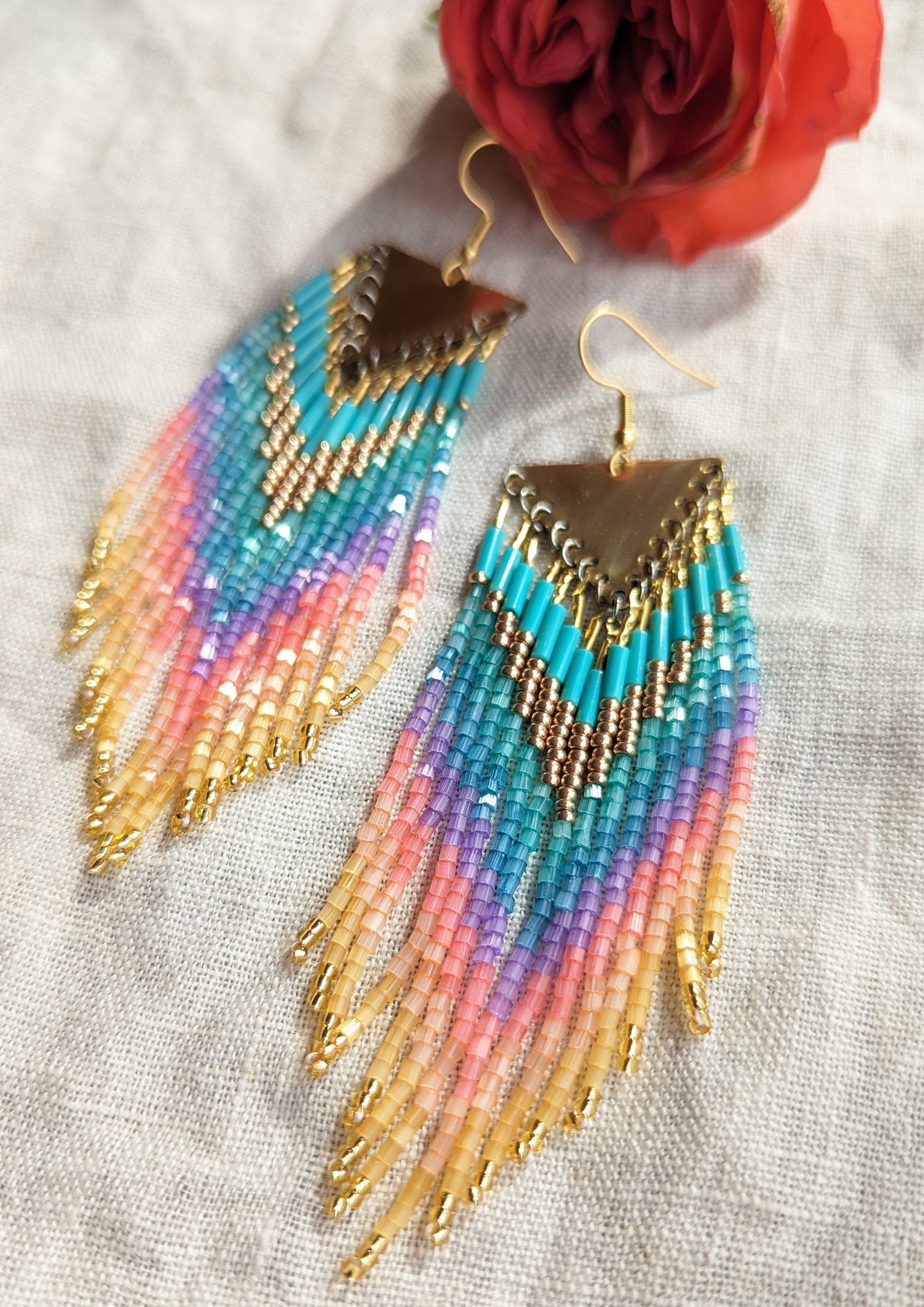 Moon & Milk beaded earrings with brass triangle tops, featuring soft rainbow tones in turquoise, purple, pink, and gold, displayed beside a red rose.