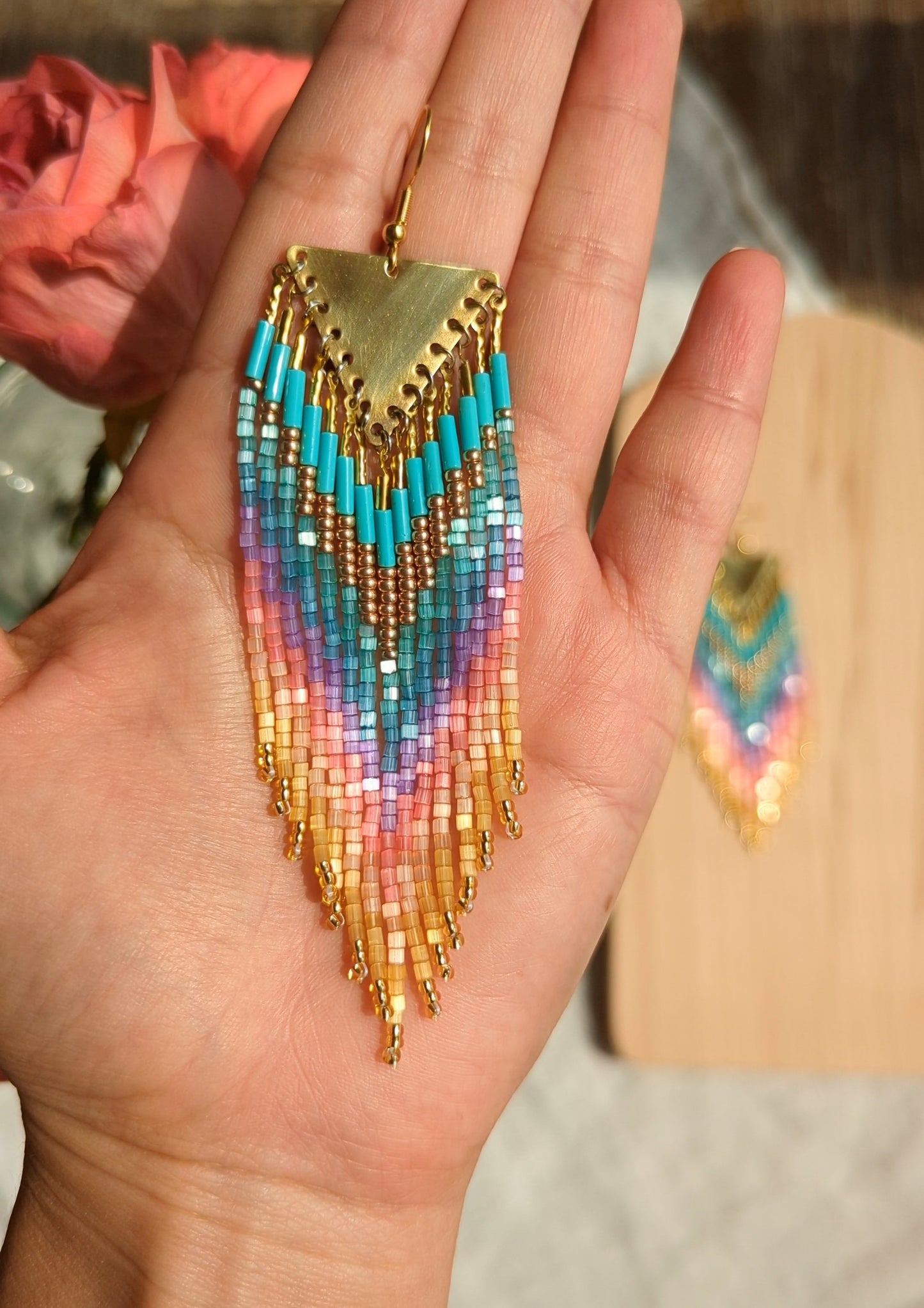 Moon & Milk beaded earrings with brass triangle tops, featuring soft rainbow tones in turquoise, purple, pink, and gold, displayed beside a red rose.