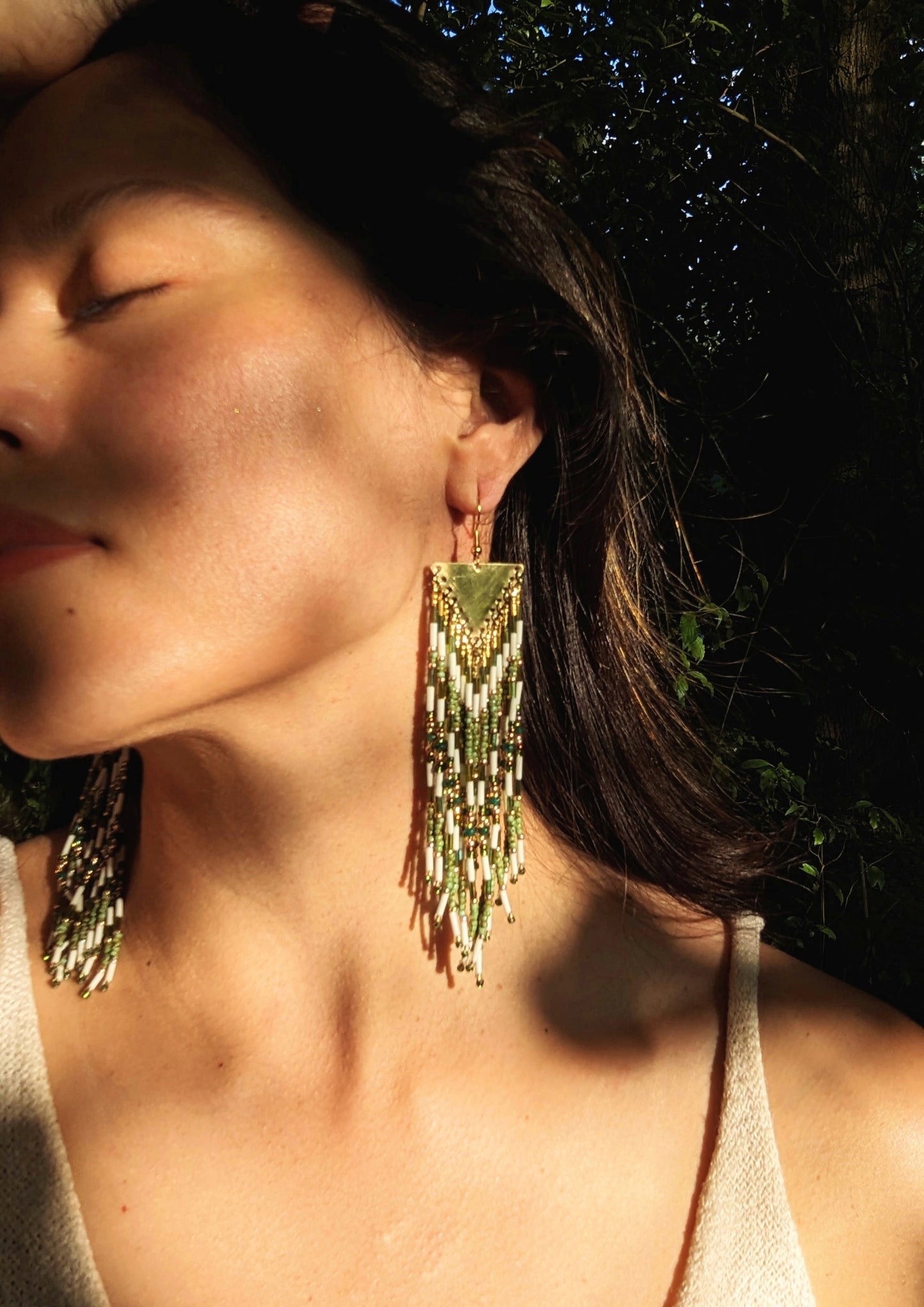 Nature-inspired long beaded earrings by Moon & Milk with green, ivory, and gold fringe, brass triangle tops, and intricate artisan beadwork worn by a model.