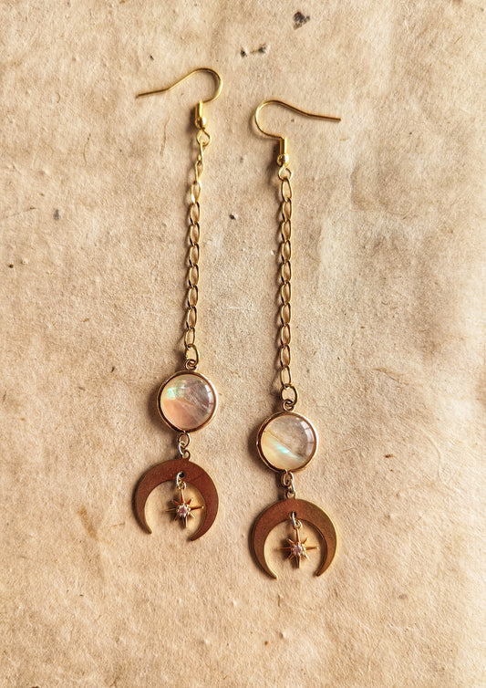 Gold chain drop earrings featuring iridescent mother of pearl accents, crescent moon charms, and delicate star details, displayed on a natural beige textured background.