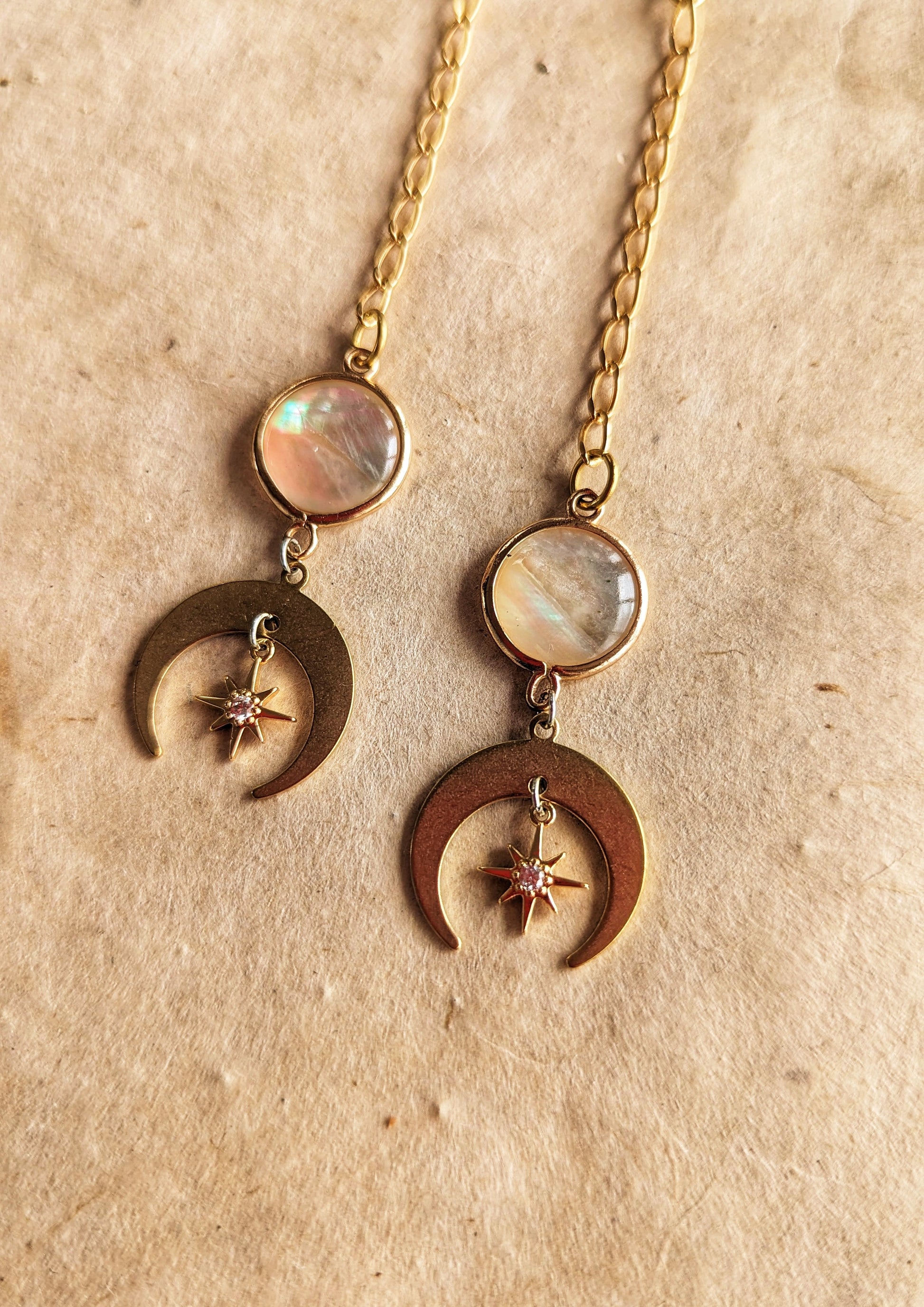 Gold chain drop earrings featuring iridescent mother of pearl accents, crescent moon charms, and delicate star details, displayed on a natural beige textured background.
