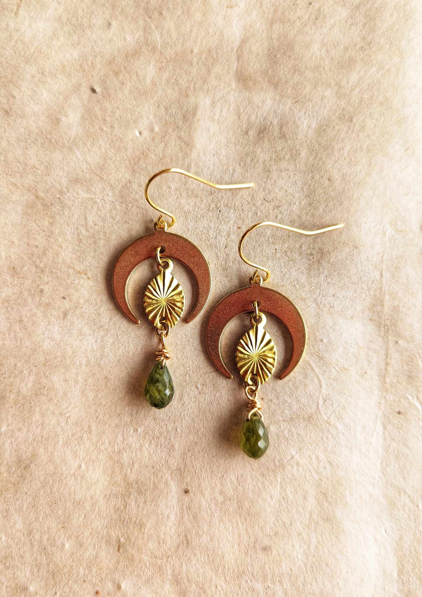 Moon & Milk -Gold brass earrings with crescent moon shapes, sunburst design, and dangling green garnet teardrop gemstones. Beige, rustic background.
