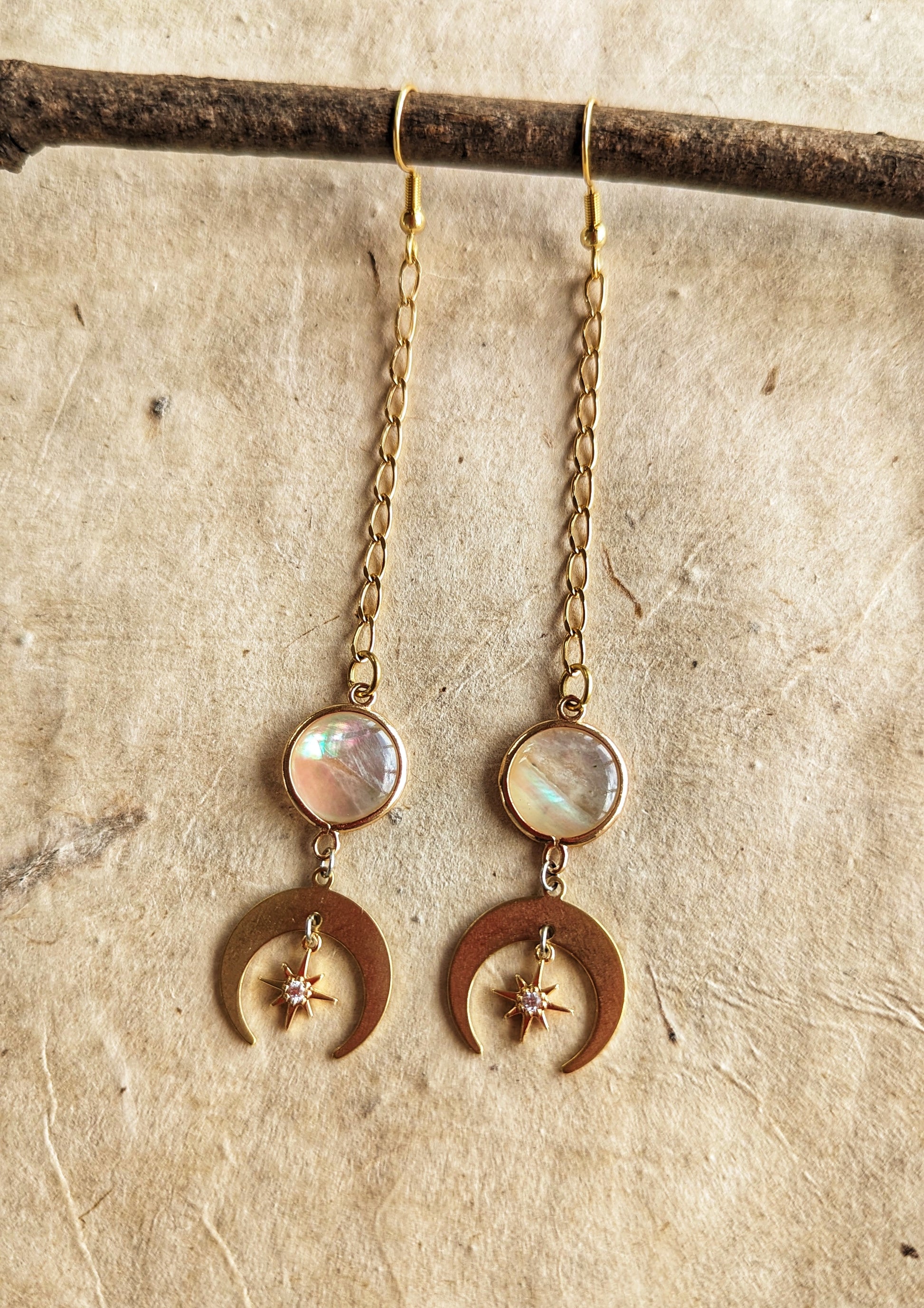 Gold chain drop earrings featuring iridescent mother of pearl accents, crescent moon charms, and delicate star details, displayed on a natural beige textured background.