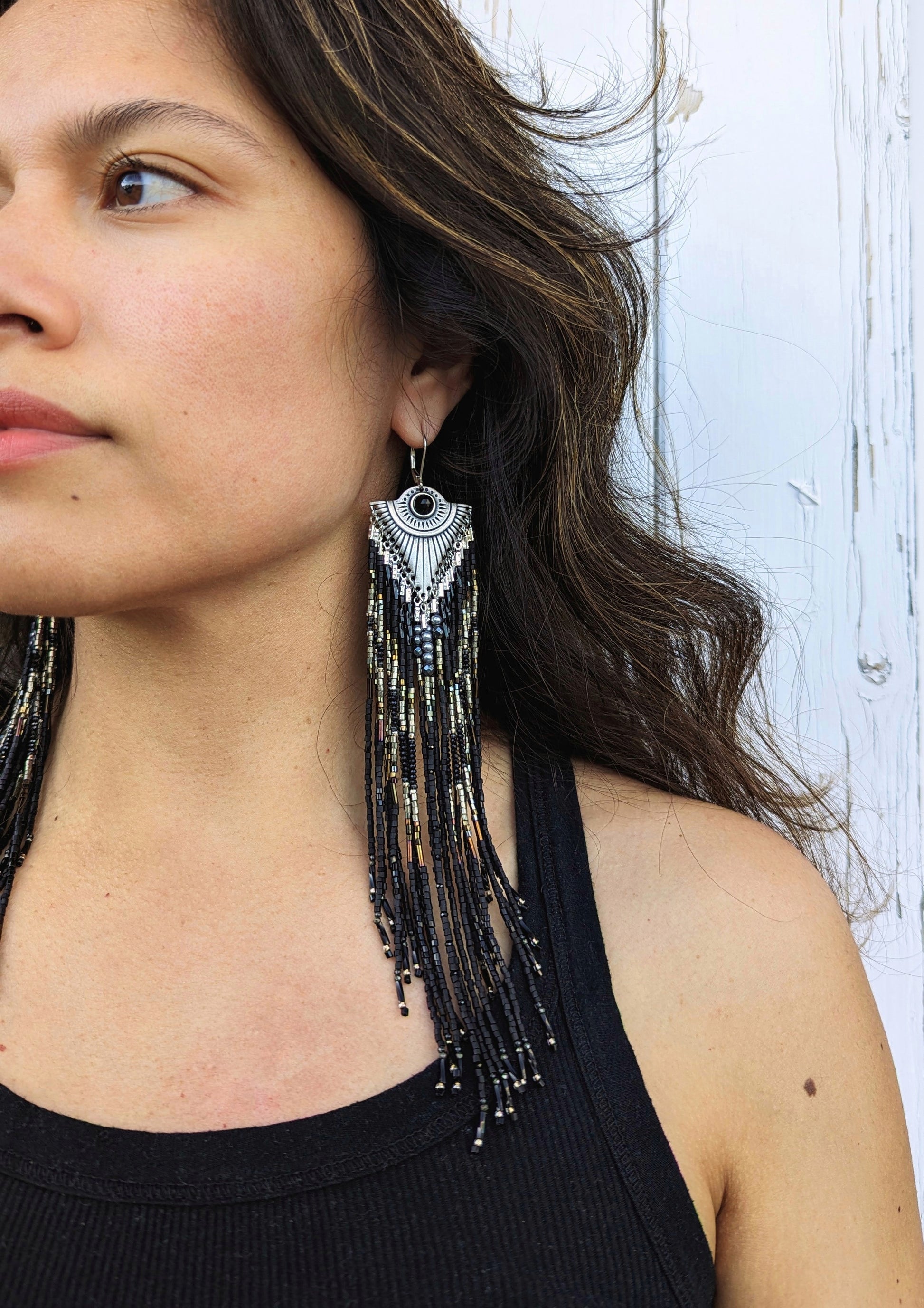 Handcrafted black beaded earrings with a fringe design on a model. The beads feature a mix of black, silver, and gold tones, complemented by a silver fan-shaped top for an elegant, bold look