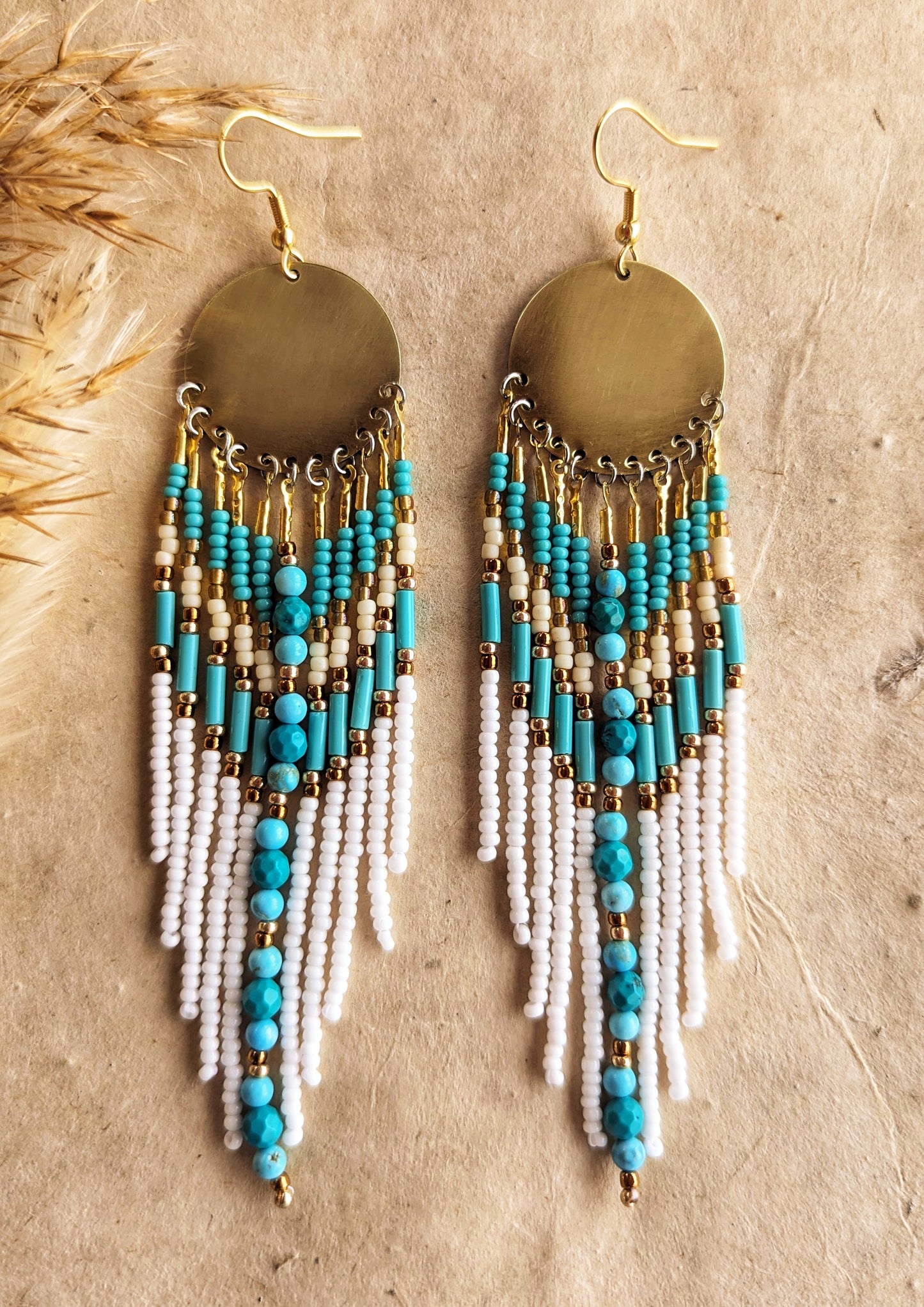 Moon & Milk earrings with turquoise stones, white fringe, brass discs, and gold accents, displayed on fabric with pampas—artistic boho seed bead jewelry.