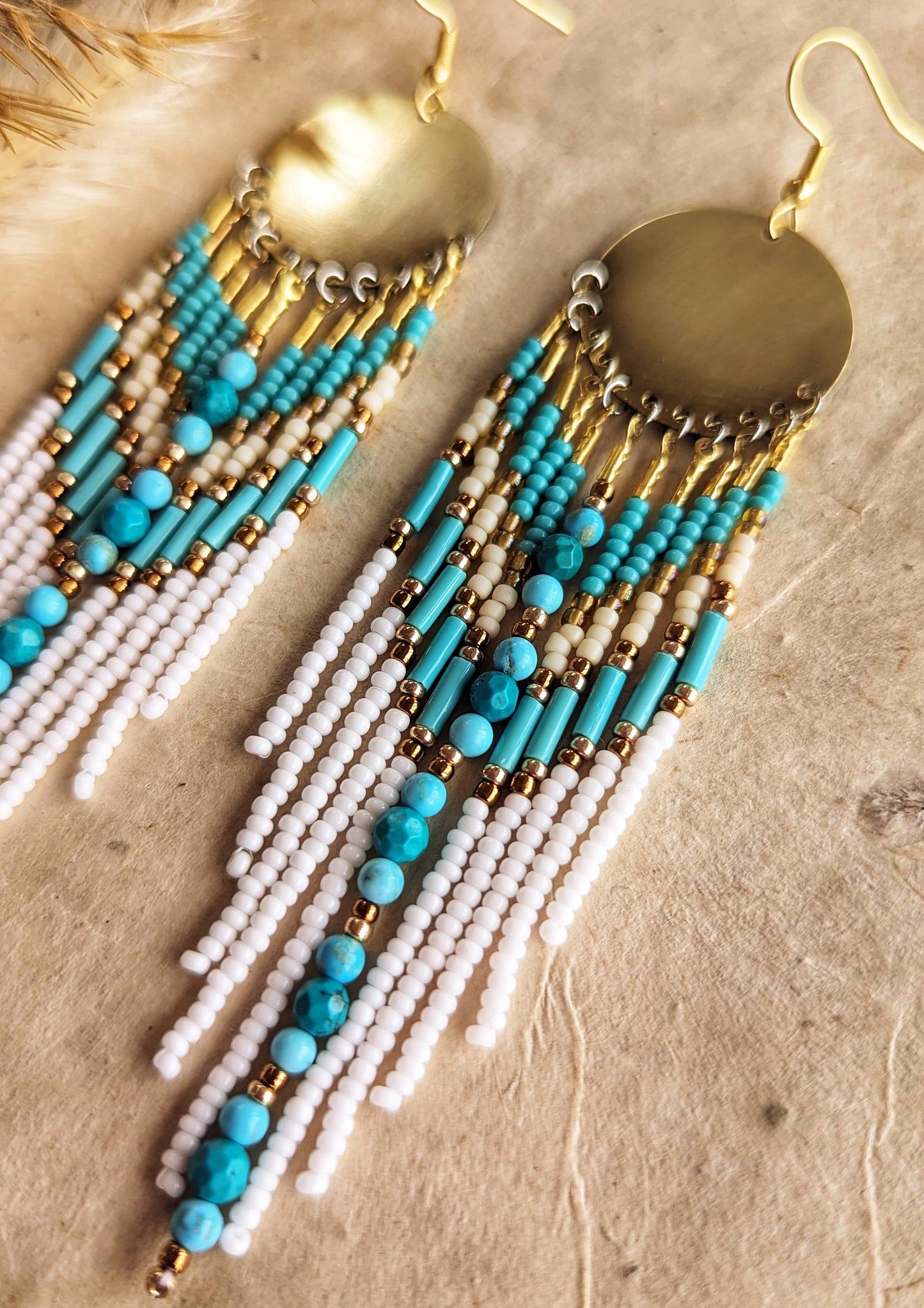 Moon & Milk earrings with turquoise stones, white fringe, brass discs, and gold accents, displayed on fabric with pampas—artistic boho seed bead jewelry.