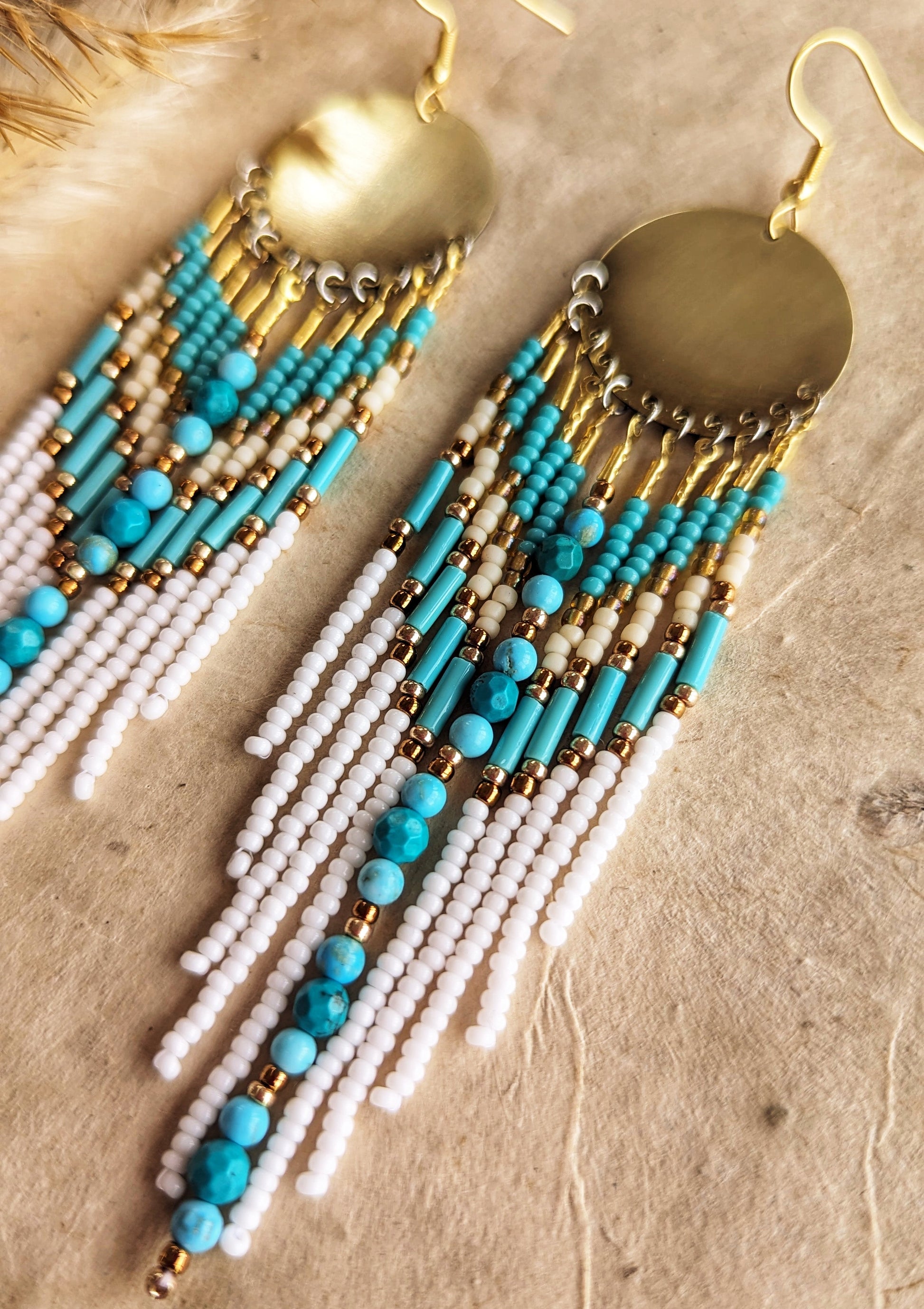 Moon & Milk earrings with turquoise stones, white fringe, brass discs, and gold accents, displayed on fabric with pampas—artistic boho seed bead jewelry.