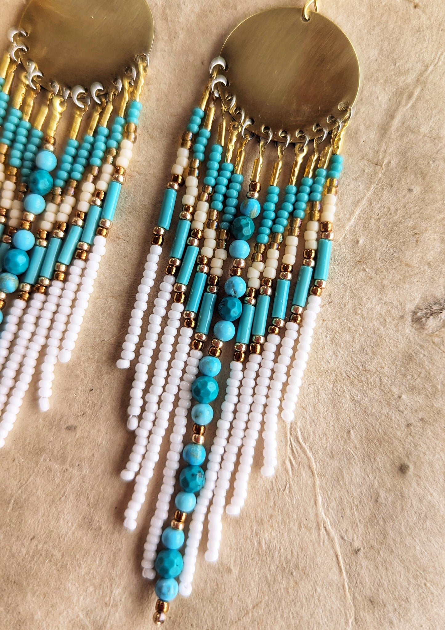Moon & Milk earrings with turquoise stones, white fringe, brass discs, and gold accents, displayed on fabric with pampas—artistic boho seed bead jewelry.