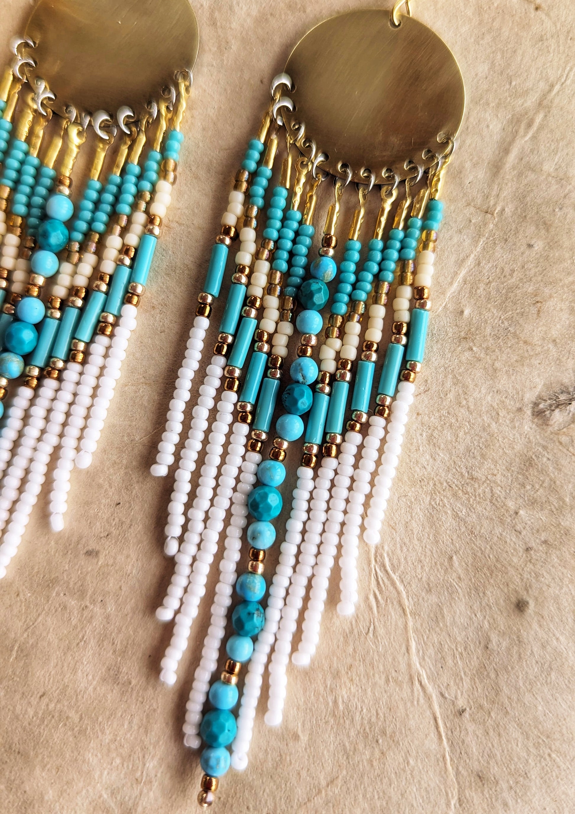 Moon & Milk earrings with turquoise stones, white fringe, brass discs, and gold accents, displayed on fabric with pampas—artistic boho seed bead jewelry.