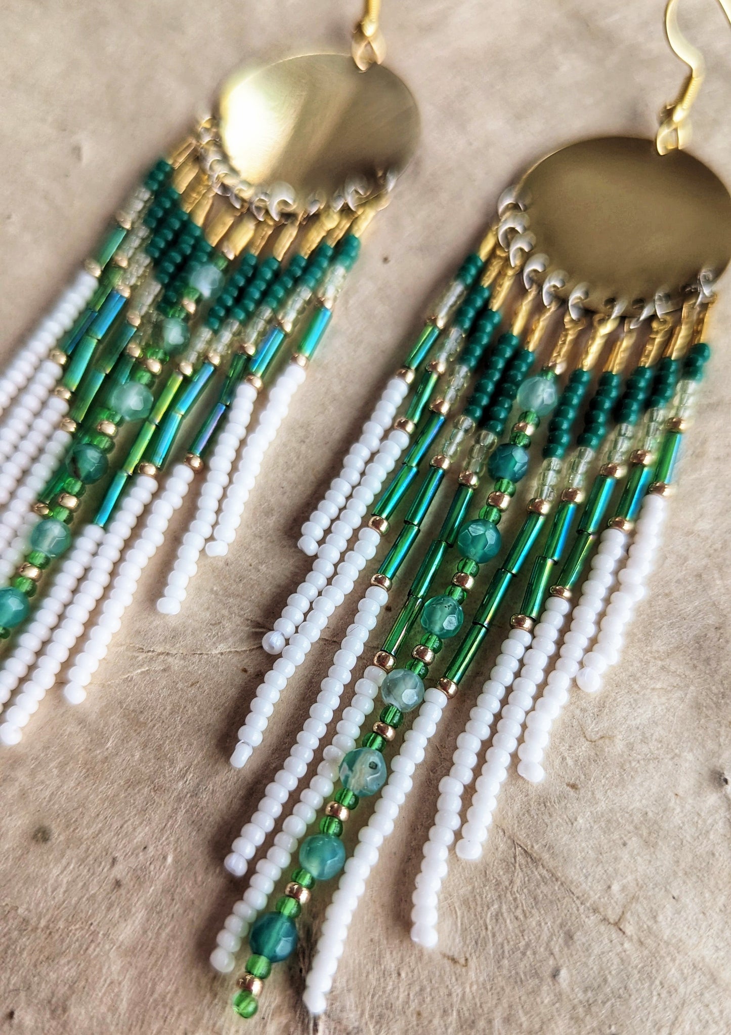 Gold dangling seed bead earrings with white and green beaded strands, featuring large circular brass discs at the top and green agate stones in the middle. Photographed on a beige, rustic background.