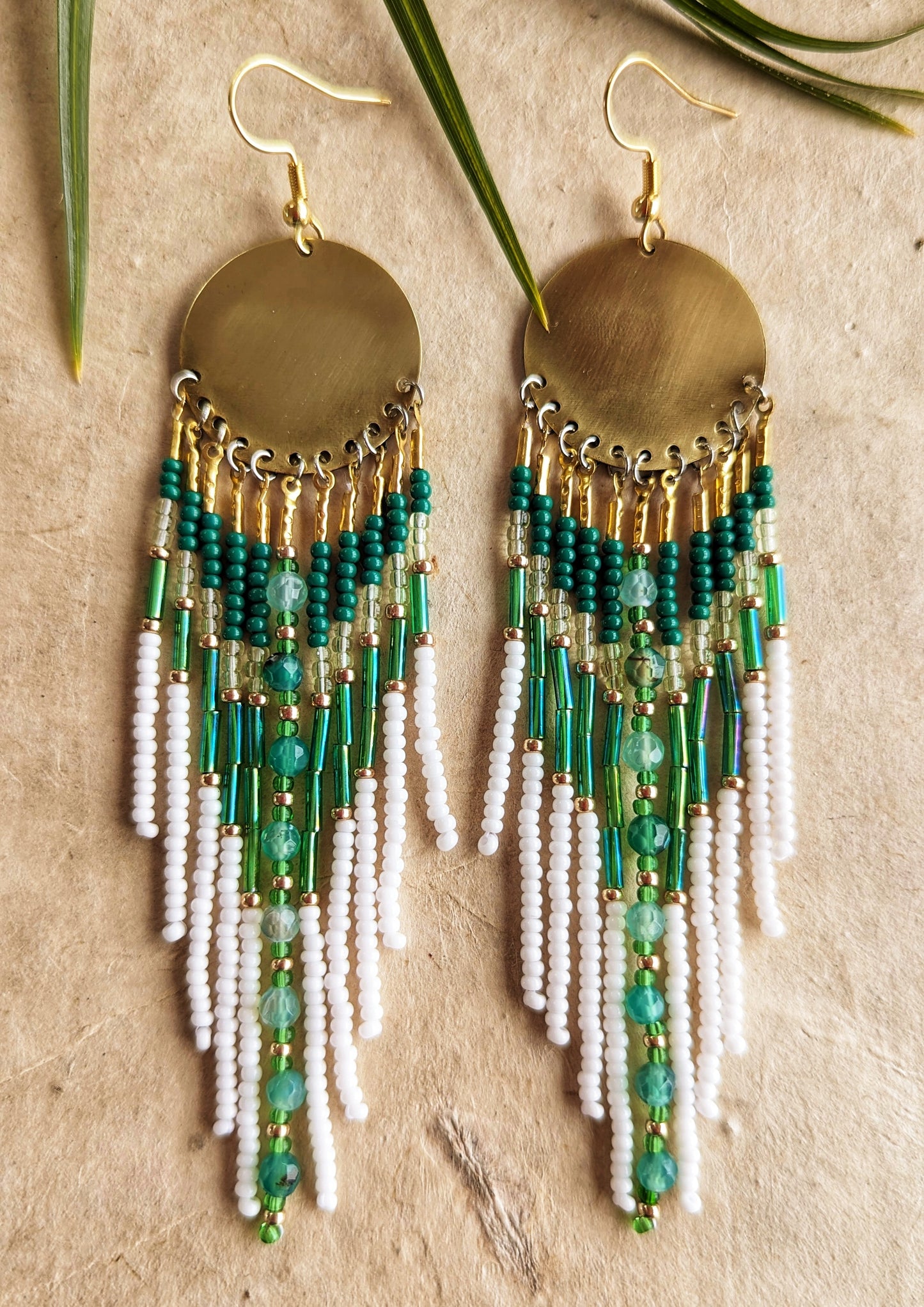 Gold dangling seed bead earrings with white and green beaded strands, featuring large circular brass discs at the top and green agate stones in the middle. Photographed on a beige, rustic background.